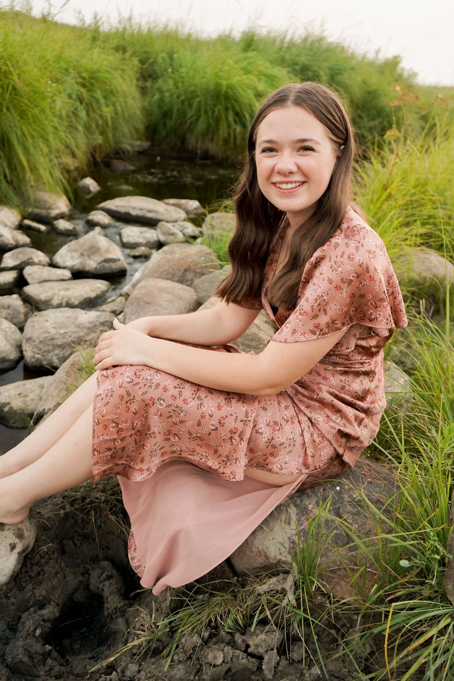 Smiling brunette teen girl in pink floral dress sitting on rocks near a stream.