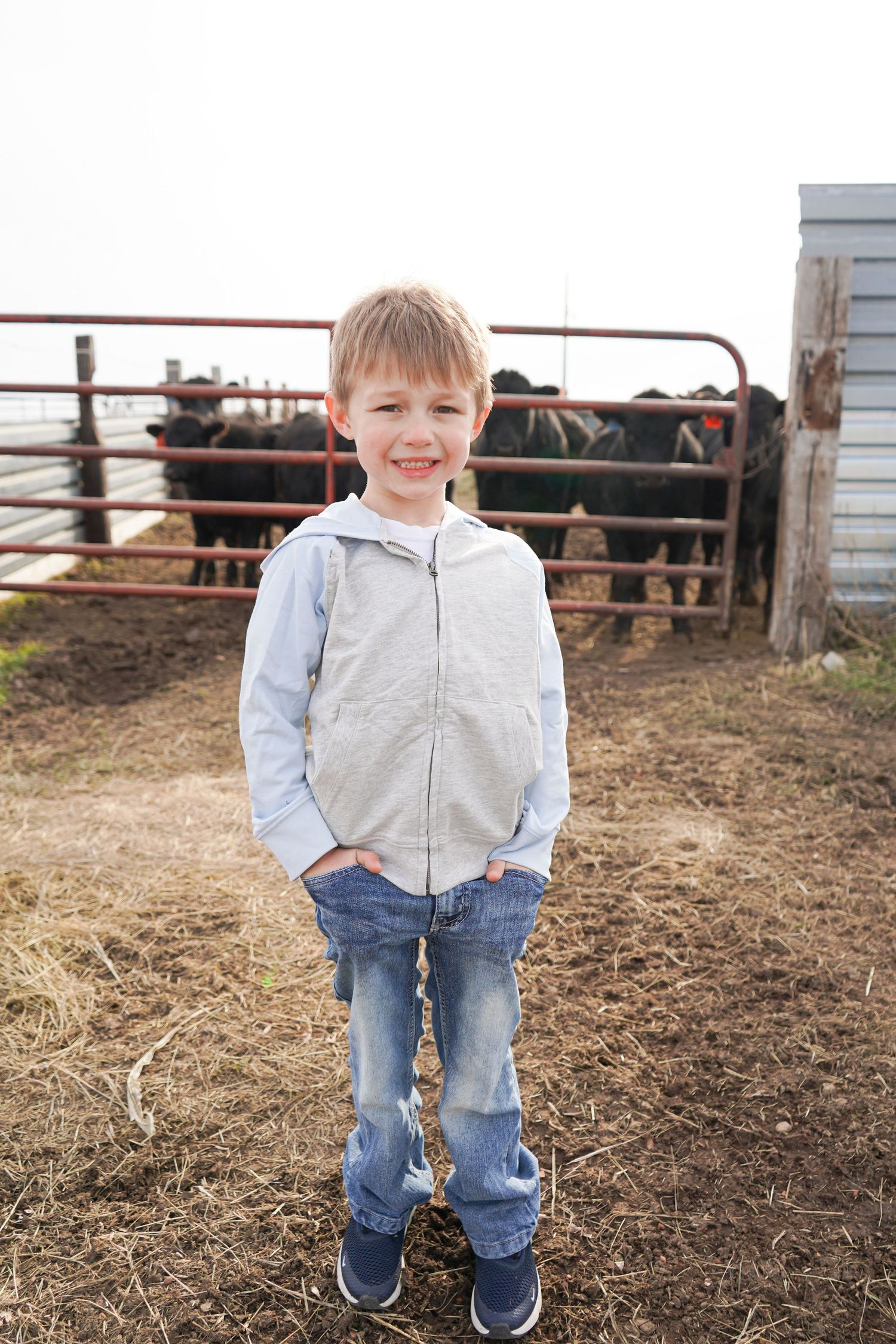 Young blonde boy in gray zip hoodie stands smiling in front of a cattle pen on a farm.