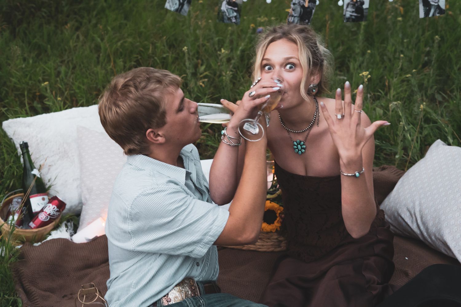 Engaged couple celebrating with champagne on a cozy outdoor picnic blanket surrounded by sunflowers.