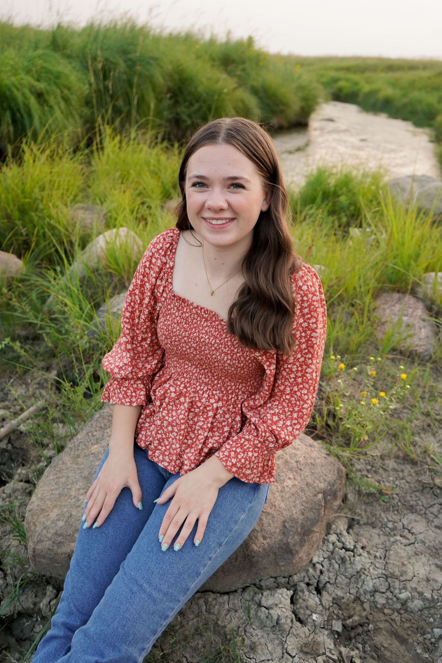Smiling brunette teen girl in red floral top sitting on rocks near a stream outdoors.