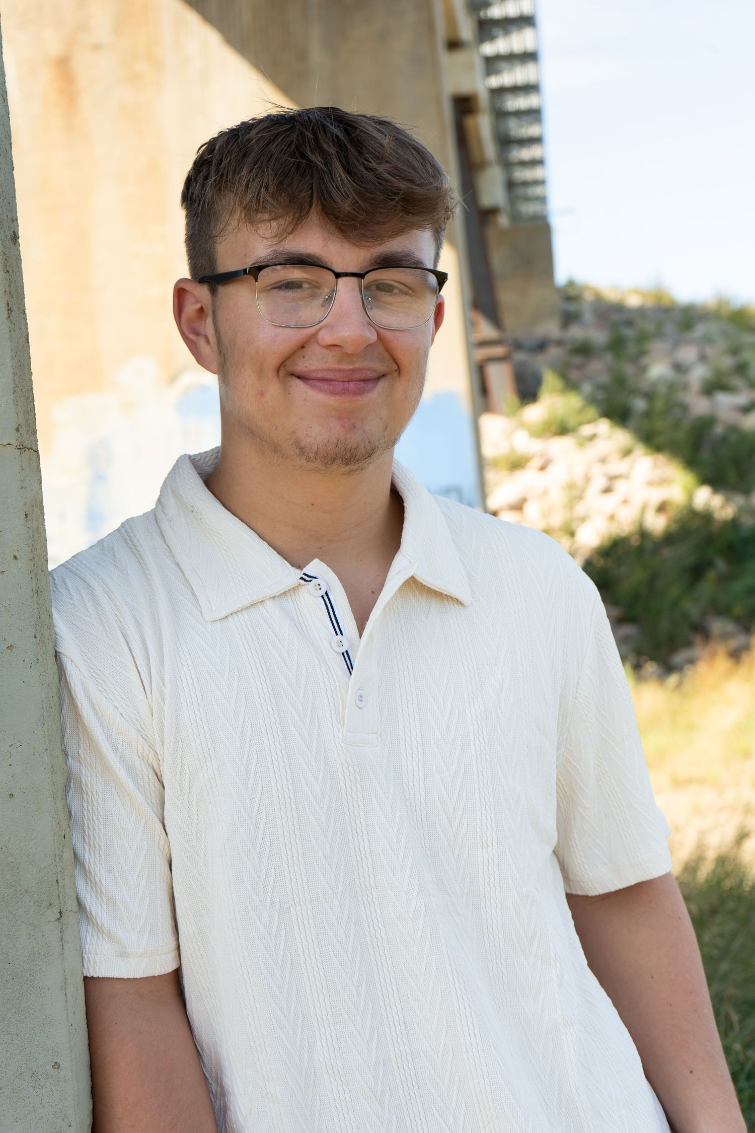 Smiling teenage boy wearing glasses and white textured polo shirt leaning against concrete pillar.