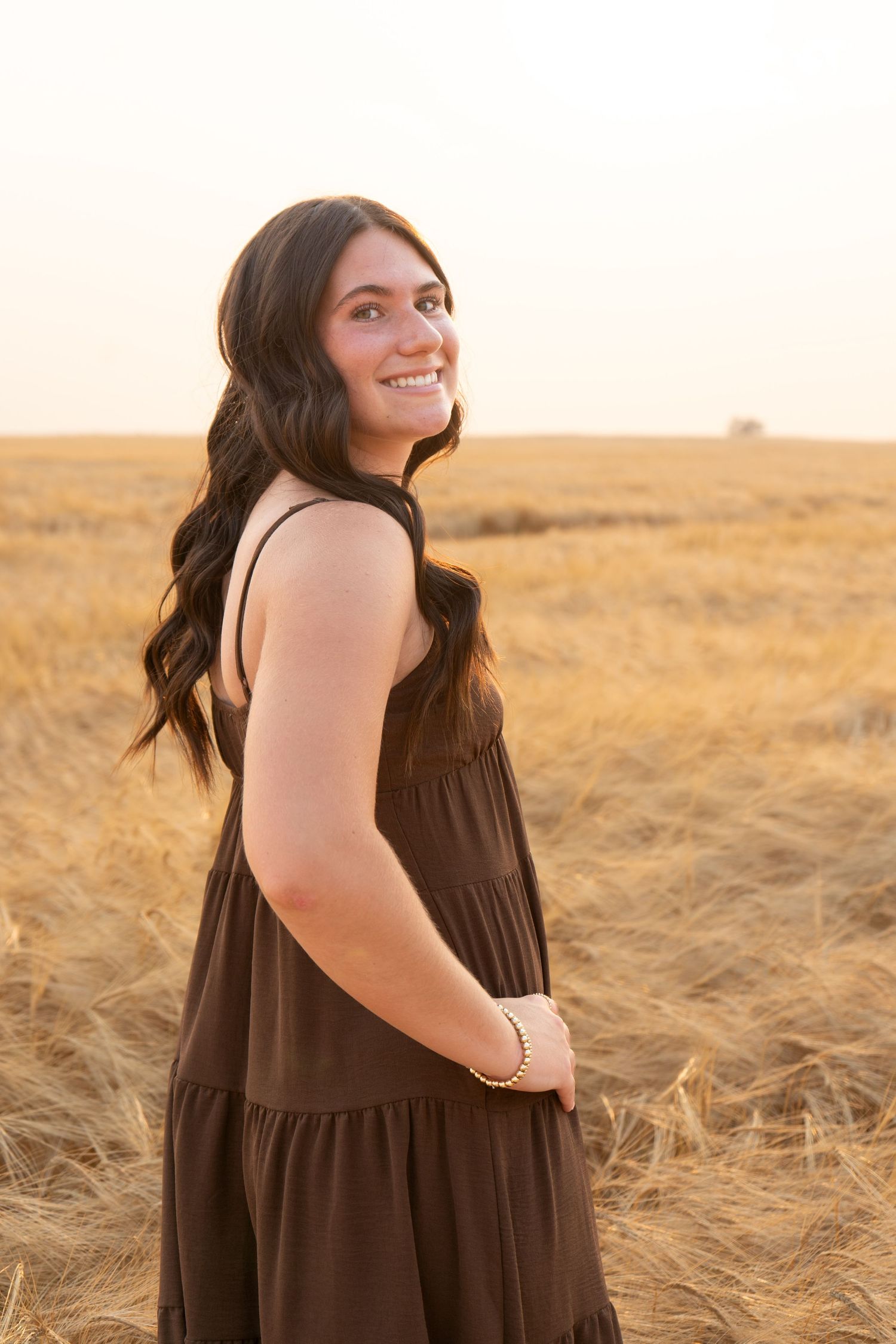 Smiling brunette teen in brown sundress stands in harvested wheat field during golden hour sunset.