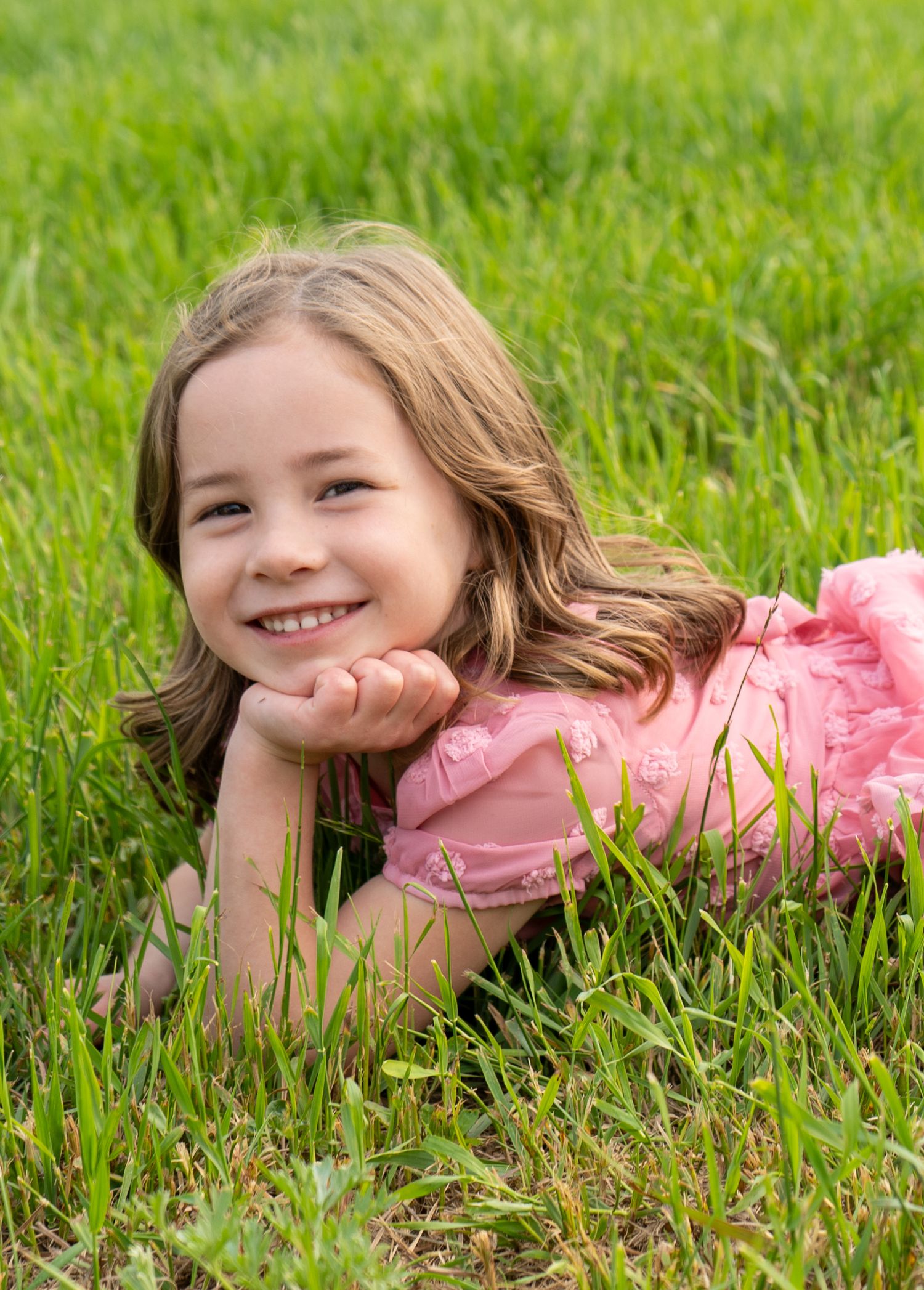 Smiling young girl in pink dress lying in green grass field.