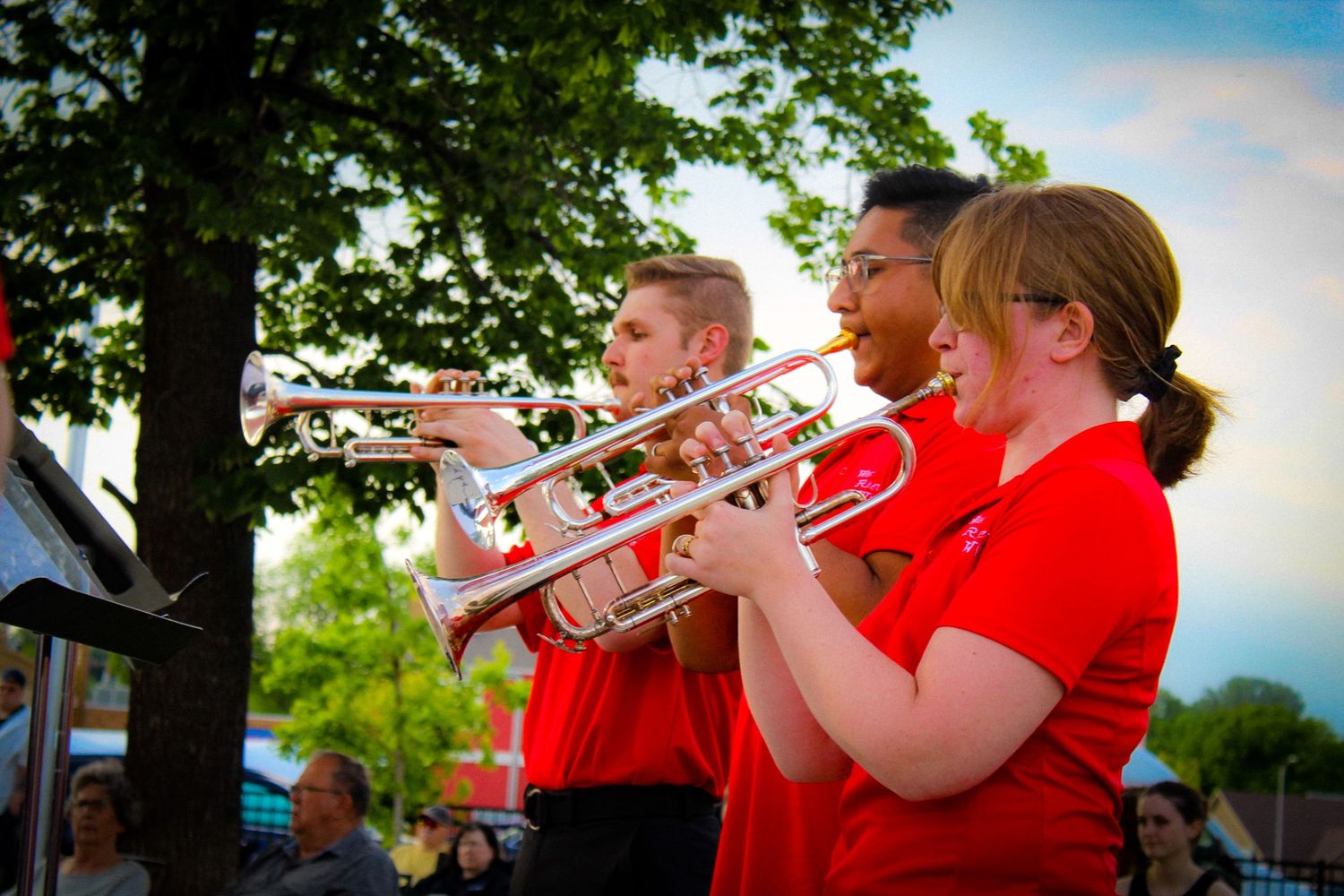 Three trumpet players in red uniforms performing at an outdoor evening band concert.