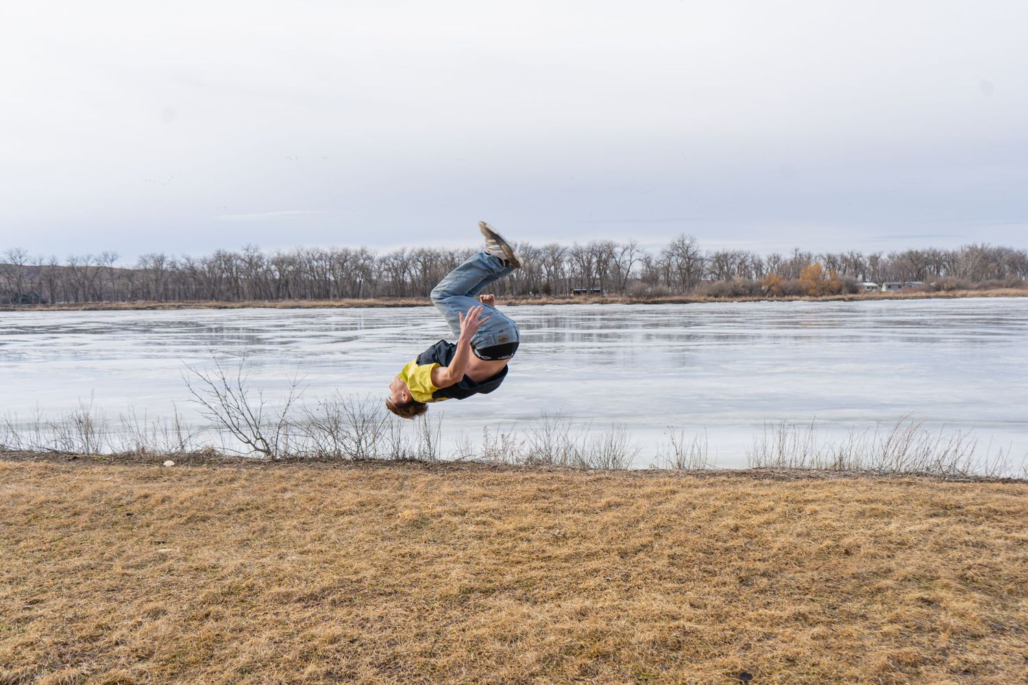 Person performing backflip near frozen river on cloudy day in grassy outdoor setting.