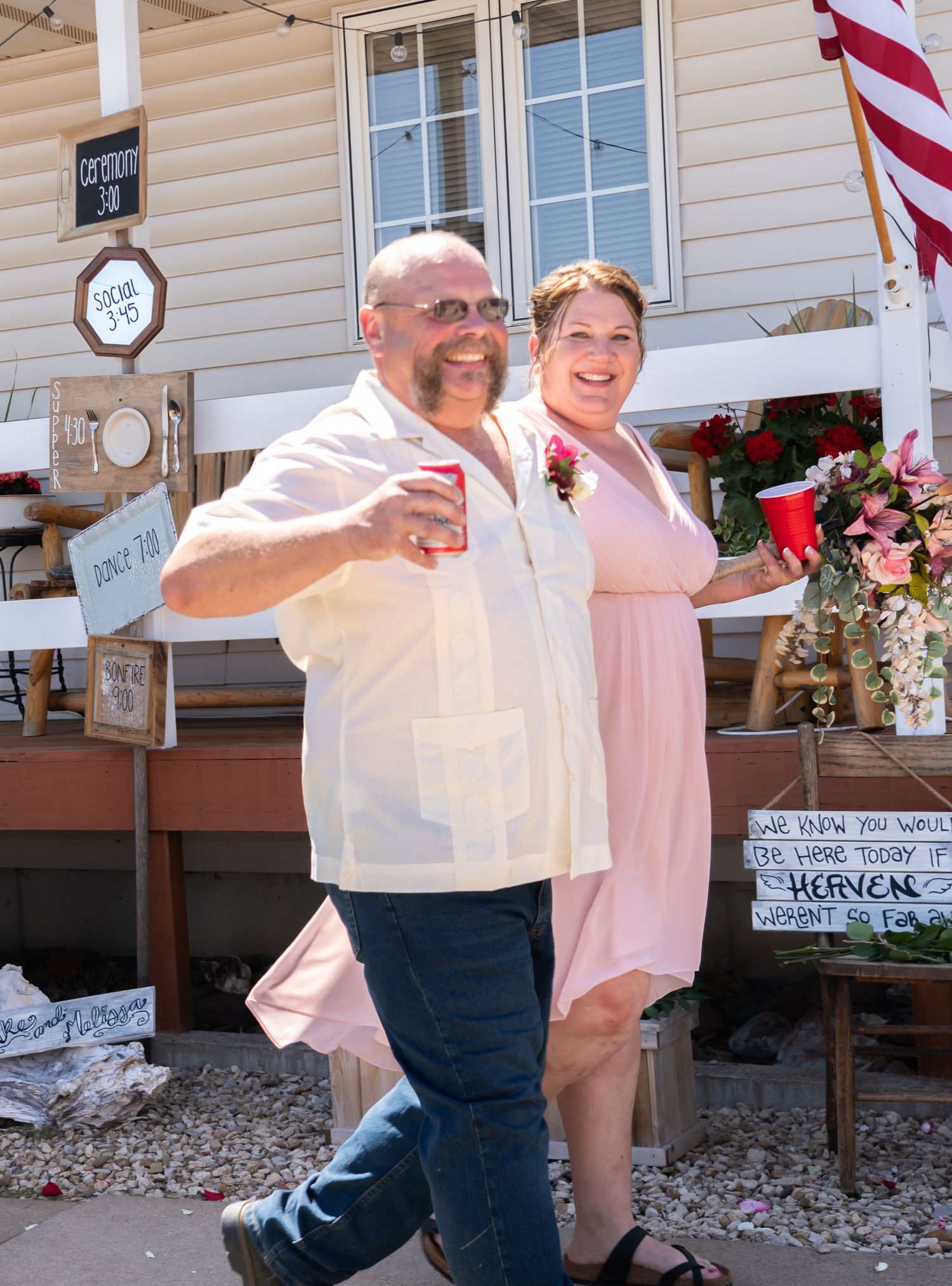 A smiling couple celebrates at an outdoor wedding reception holding red cups in front of a decorated house.