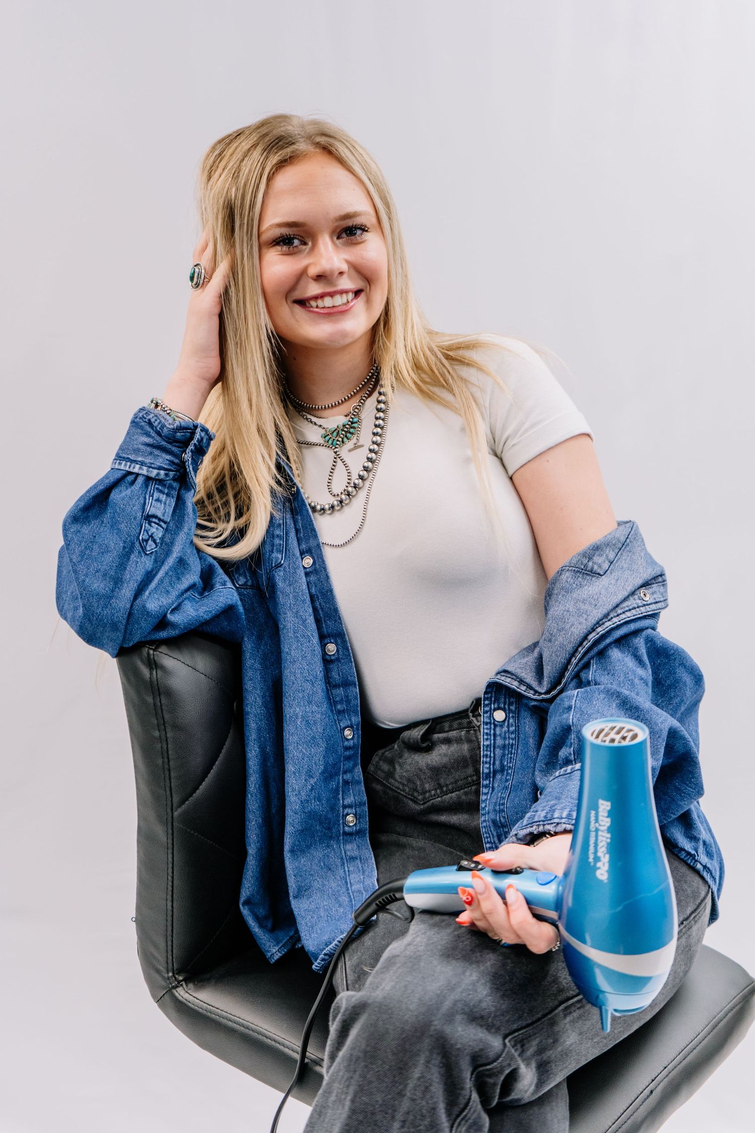 Smiling blonde woman sitting in black chair holding blue hair dryer wearing denim jacket and layered necklaces.