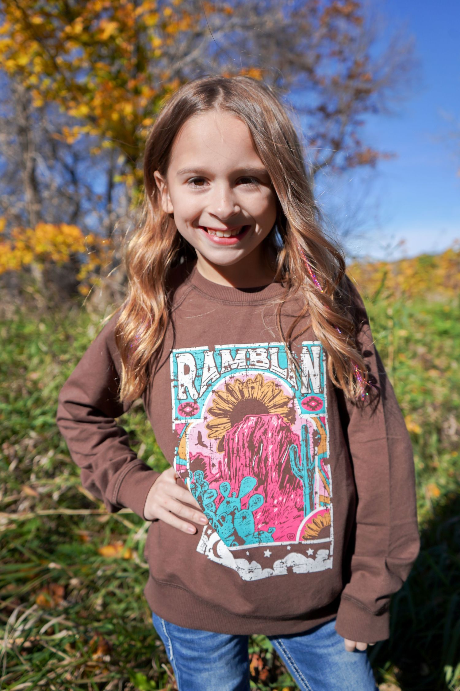 Smiling young girl in brown Ramblin graphic sweatshirt posing outdoors in autumn.