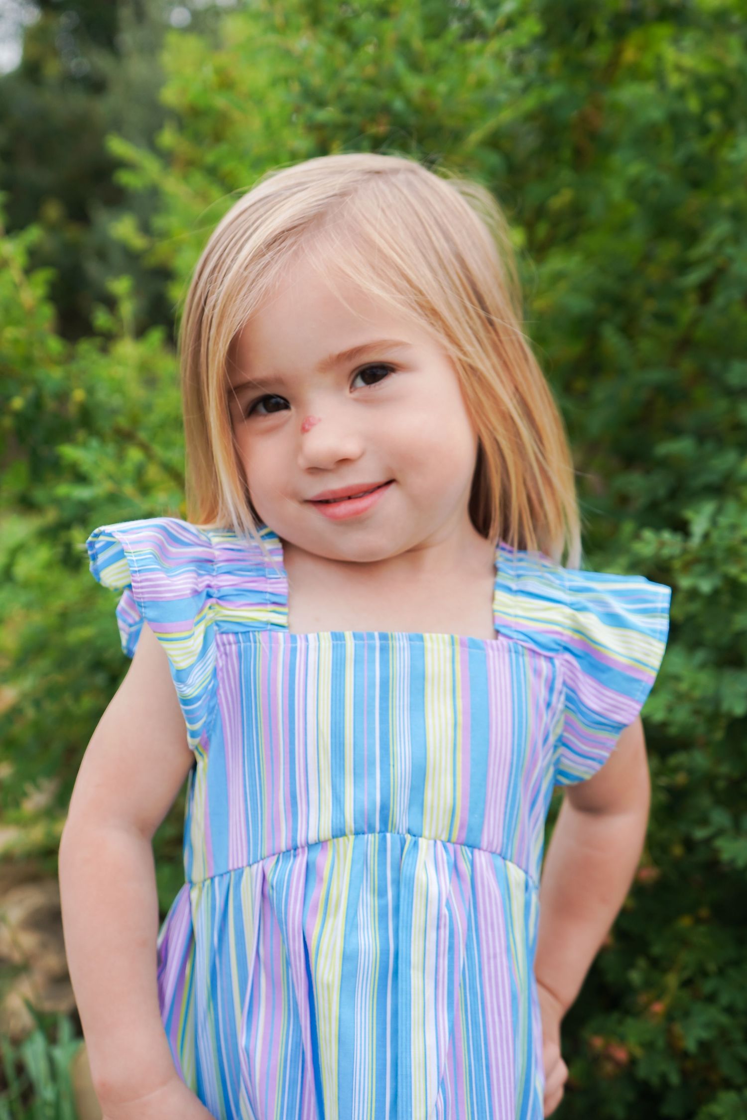 Sweet blonde toddler girl in a colorful striped dress smiling in front of green bushes outdoors.