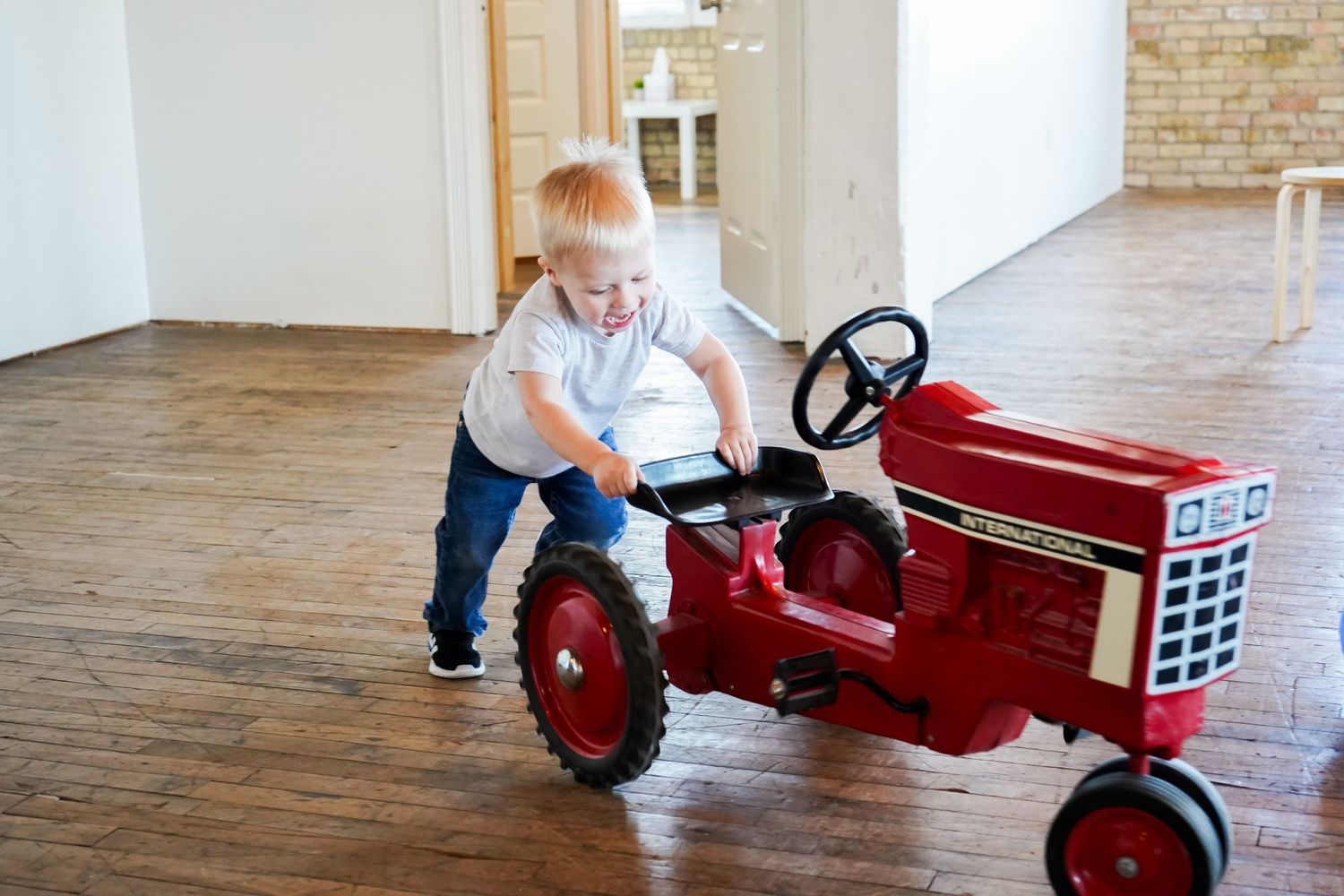 Happy toddler boy plays with a classic red International Harvester pedal tractor on hardwood floors.
