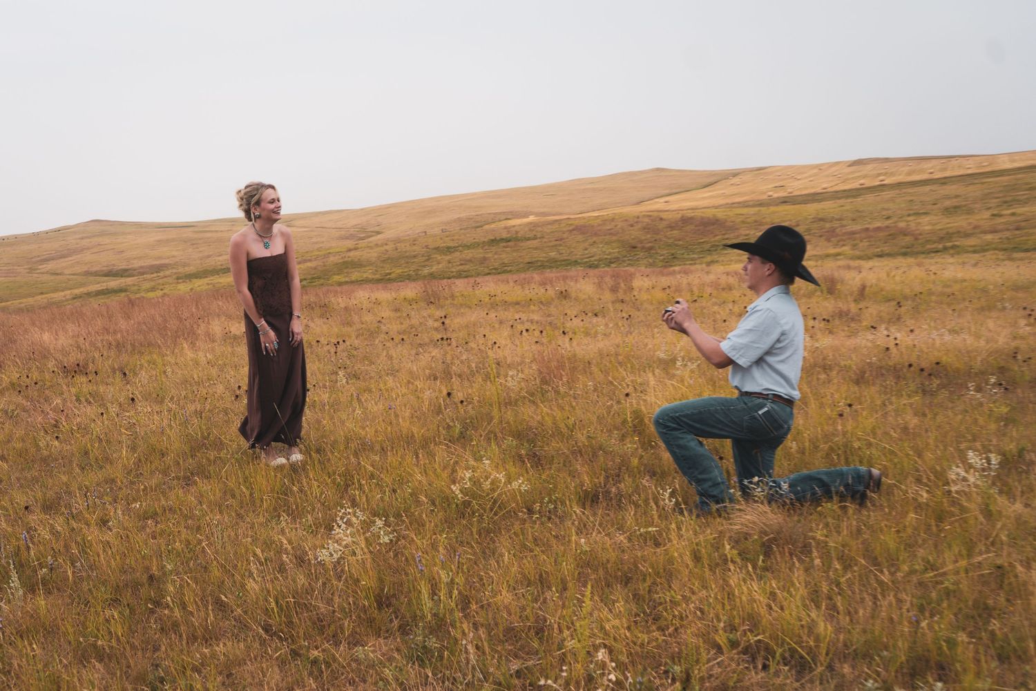 Man kneeling in golden prairie field proposing to woman in brown maxi dress during sunset.