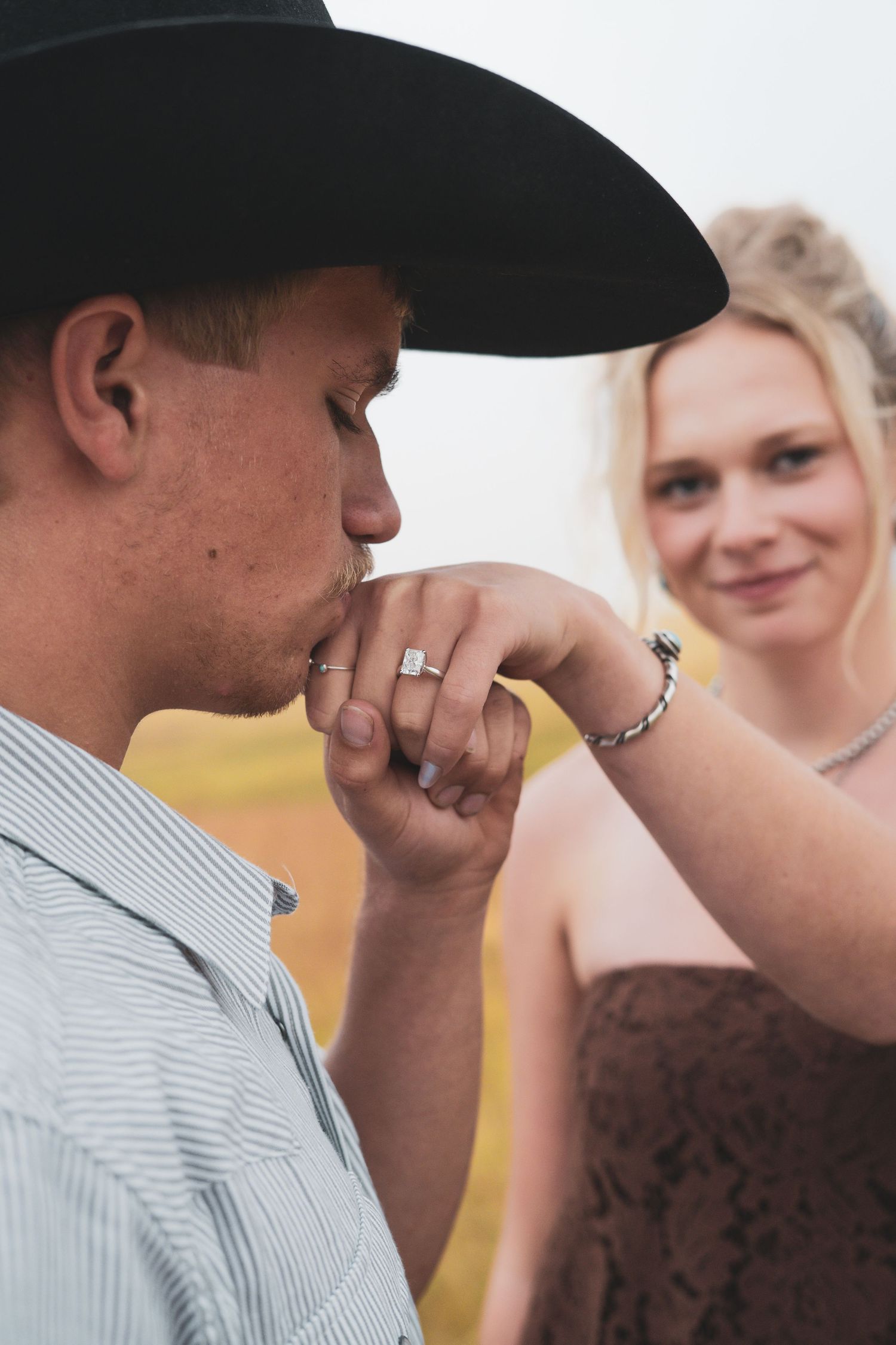 A man in a cowboy hat kisses a woman's hand displaying a diamond engagement ring as she smiles behind him.