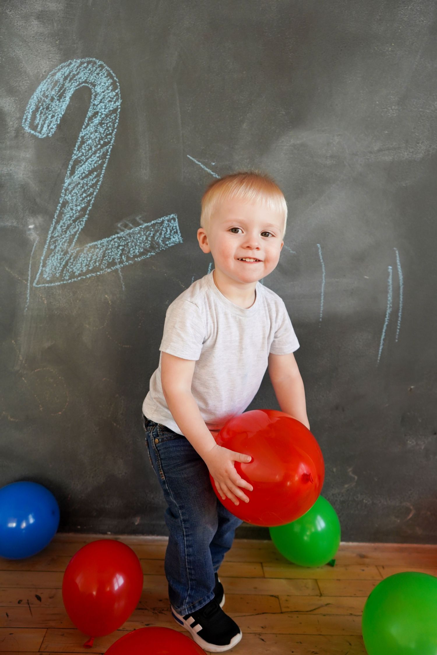 Smiling toddler boy holding a red balloon in front of a chalkboard with the number 2 drawn on it.