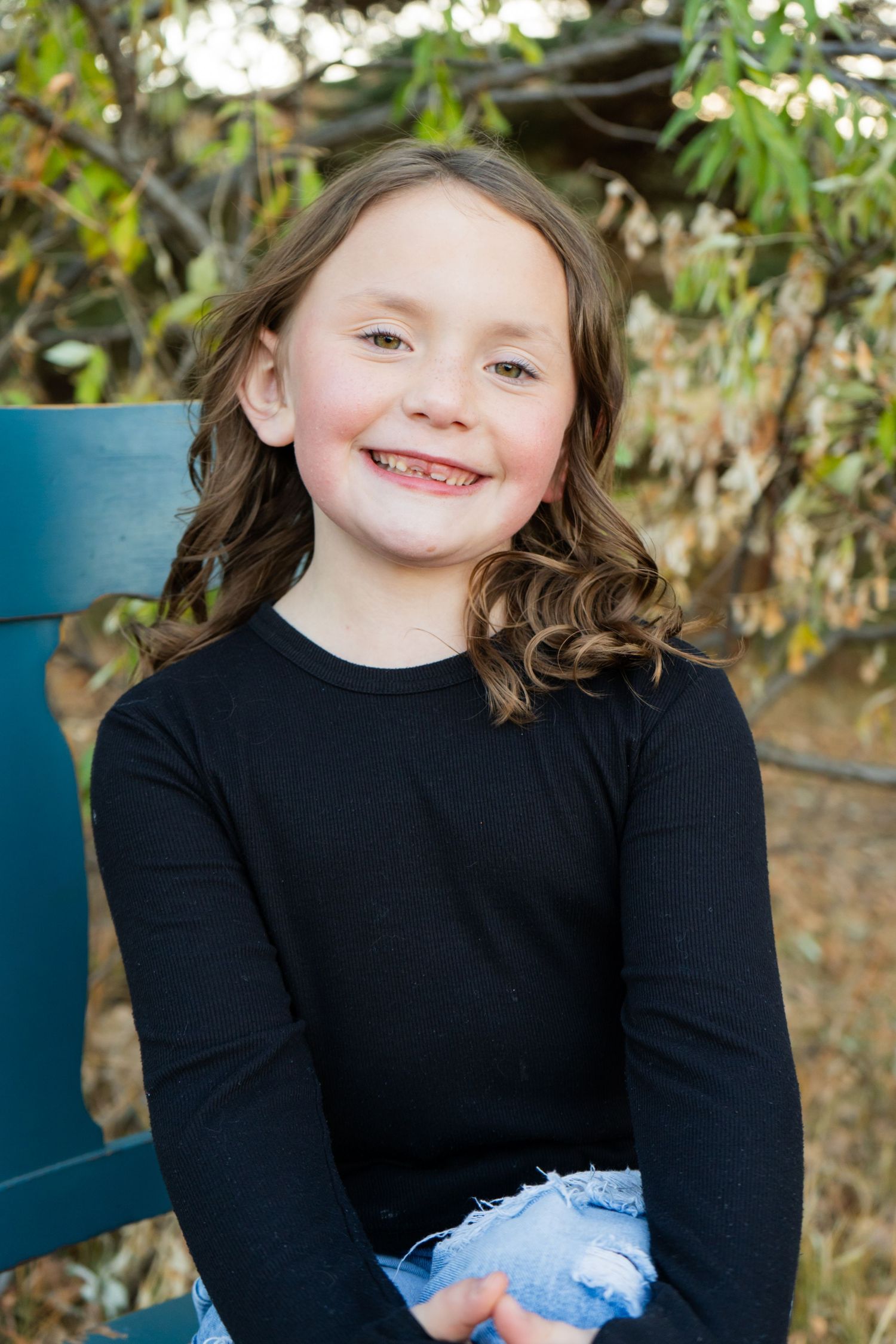 Young girl with brown curly hair smiling in black long-sleeve shirt seated on teal chair.