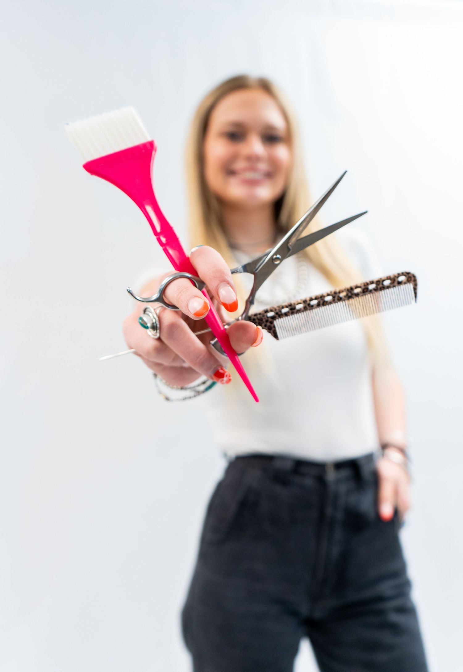 Smiling blonde hairstylist holding scissors, comb, and pink dye brush toward camera.