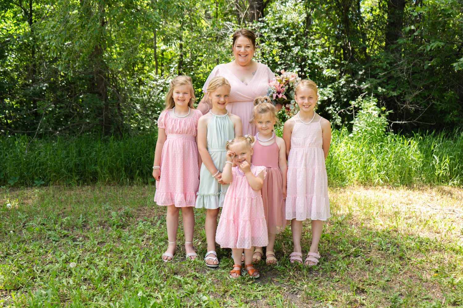 A woman poses with five young girls in pastel dresses in a sunny green outdoor setting.