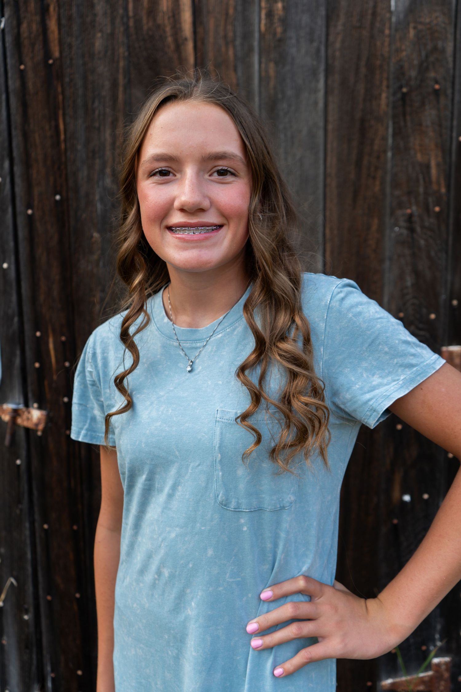 Smiling teenage girl with wavy brown hair and braces wearing a light blue t-shirt dress in front of a wooden wall.