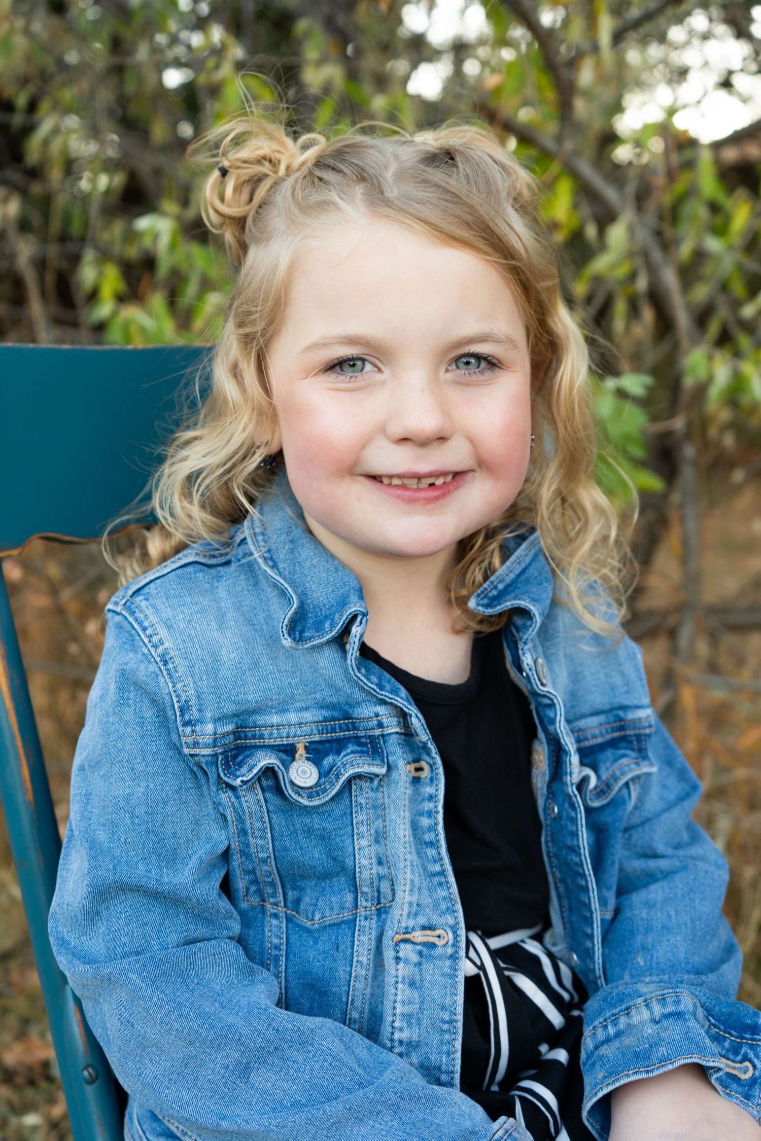 Adorable blonde curly-haired girl smiling in denim jacket seated on teal chair outdoors.