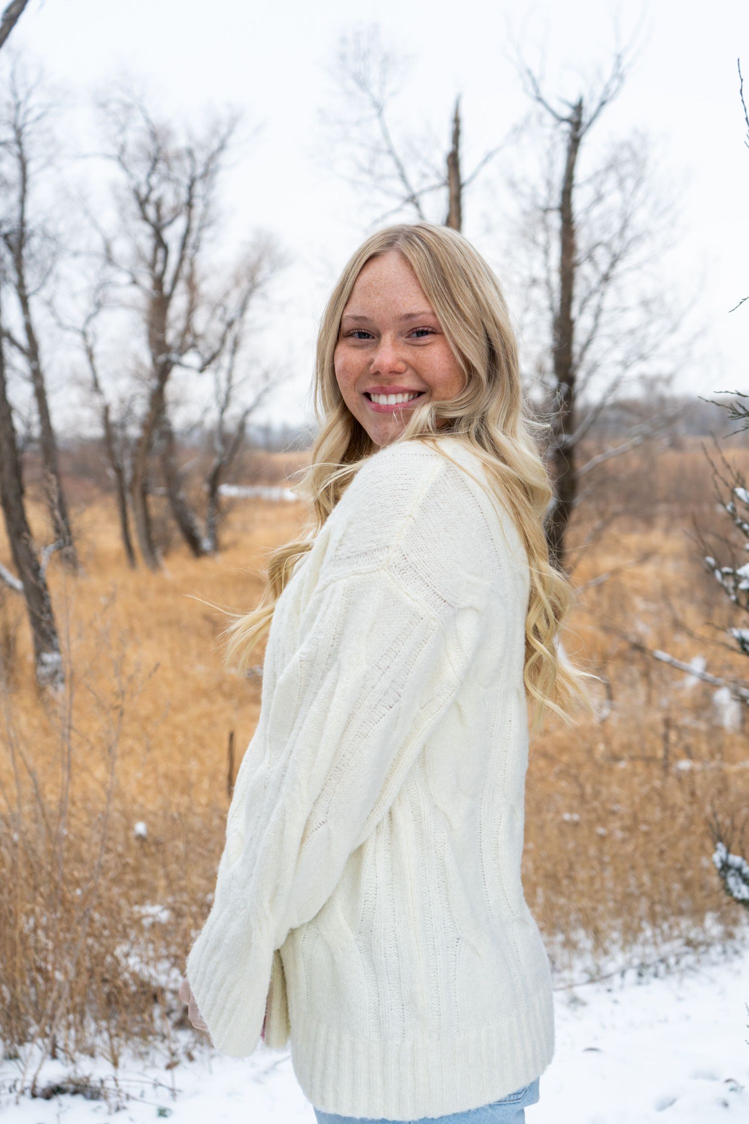 Blonde woman in cream knit sweater smiling over shoulder in snowy outdoor setting.
