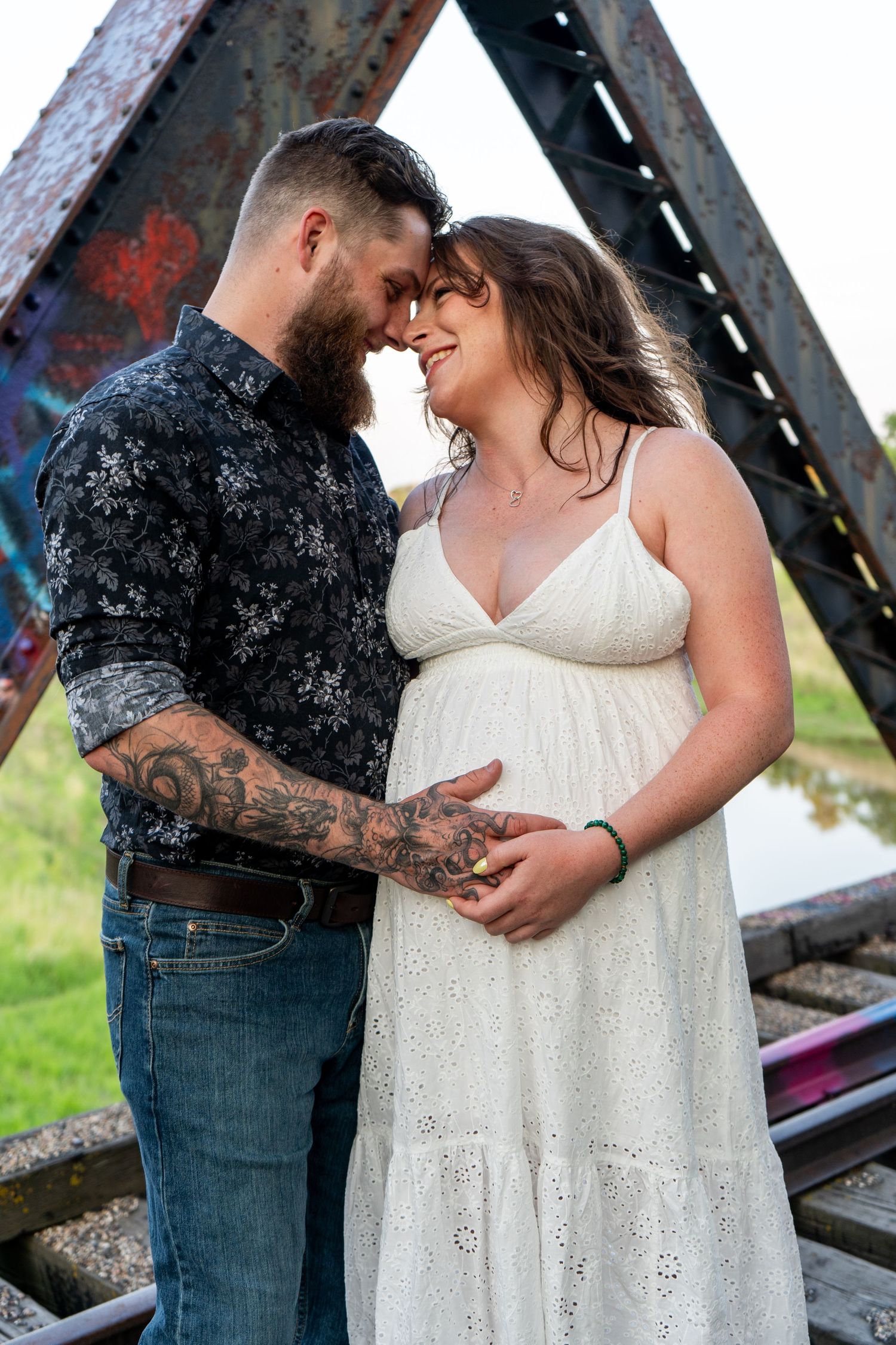 Tattooed couple touching foreheads while cradling baby bump near rustic iron bridge outdoors.