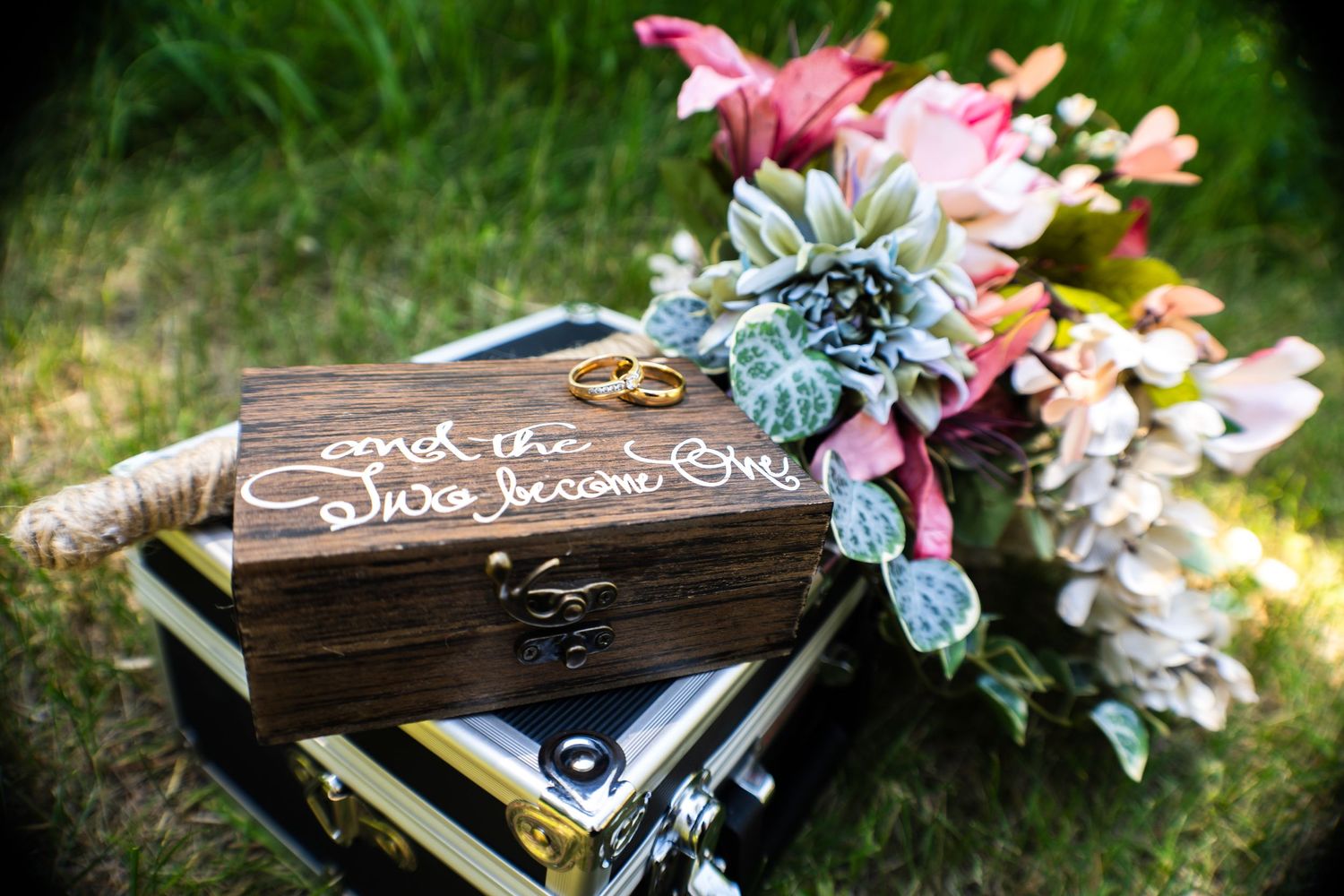 Two gold wedding rings rest atop a rustic engraved wooden box beside a colorful bridal bouquet outdoors.