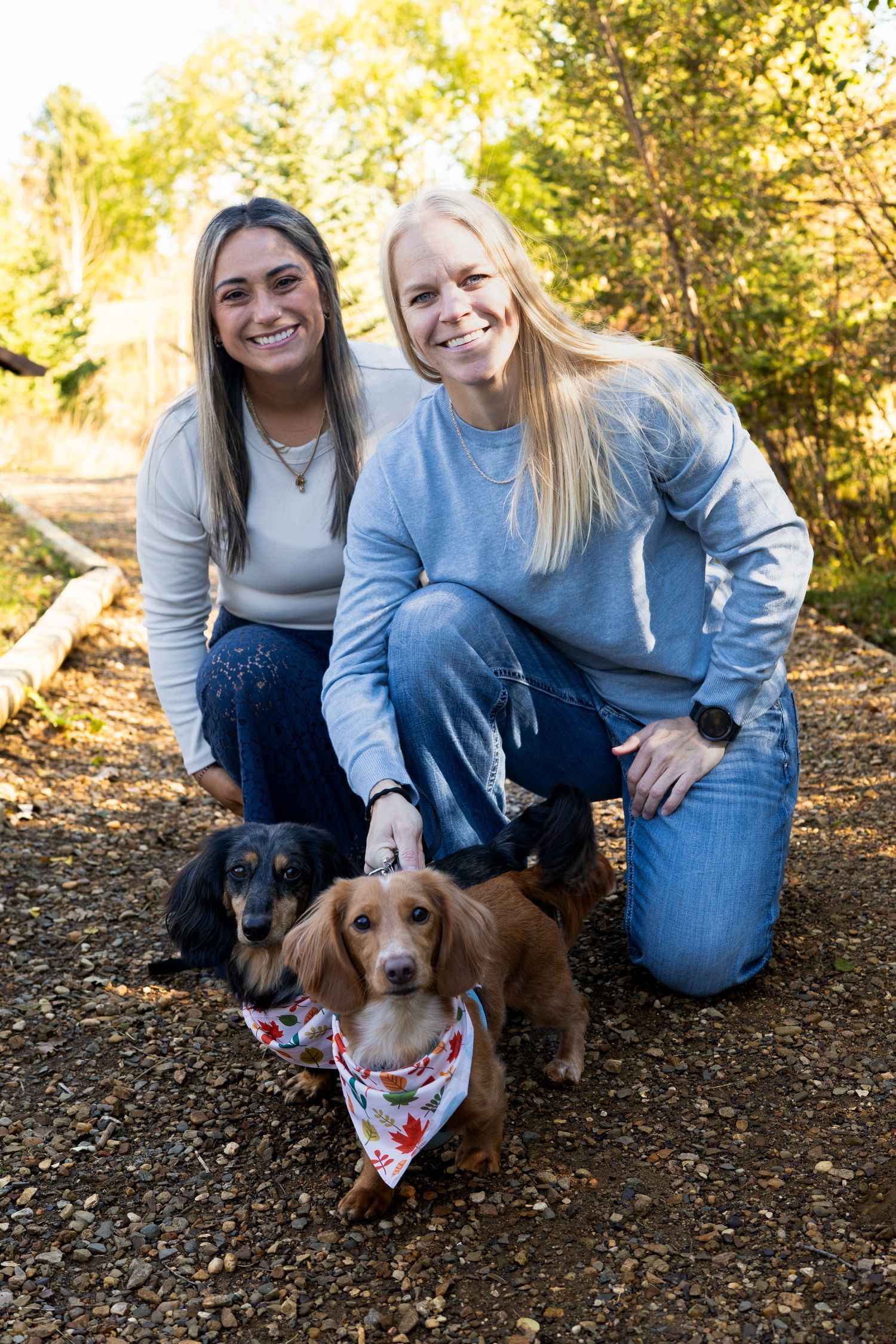 Two smiling women kneel with two dachshunds on a nature trail in autumn.