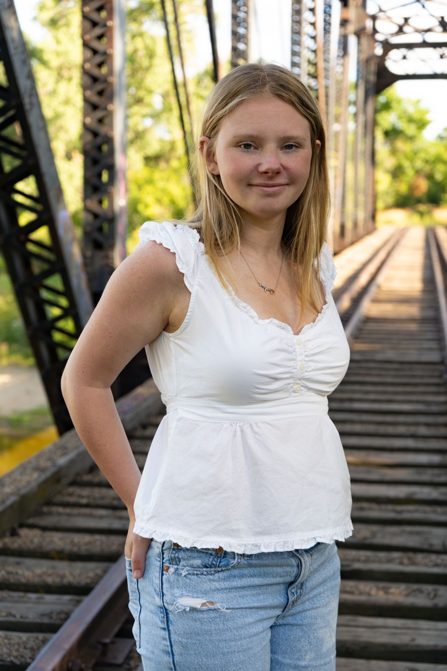 Smiling blonde teen girl stands on iron railroad bridge in white top and jeans.