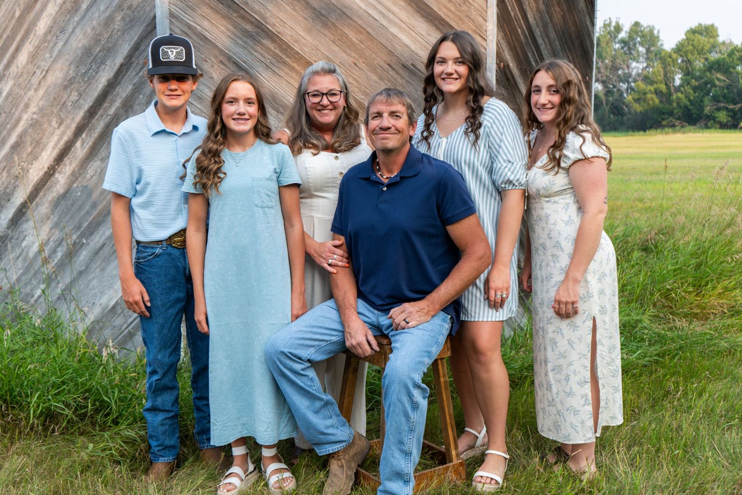 Family of six smiles together in casual attire beside a rustic wooden barn in a field.