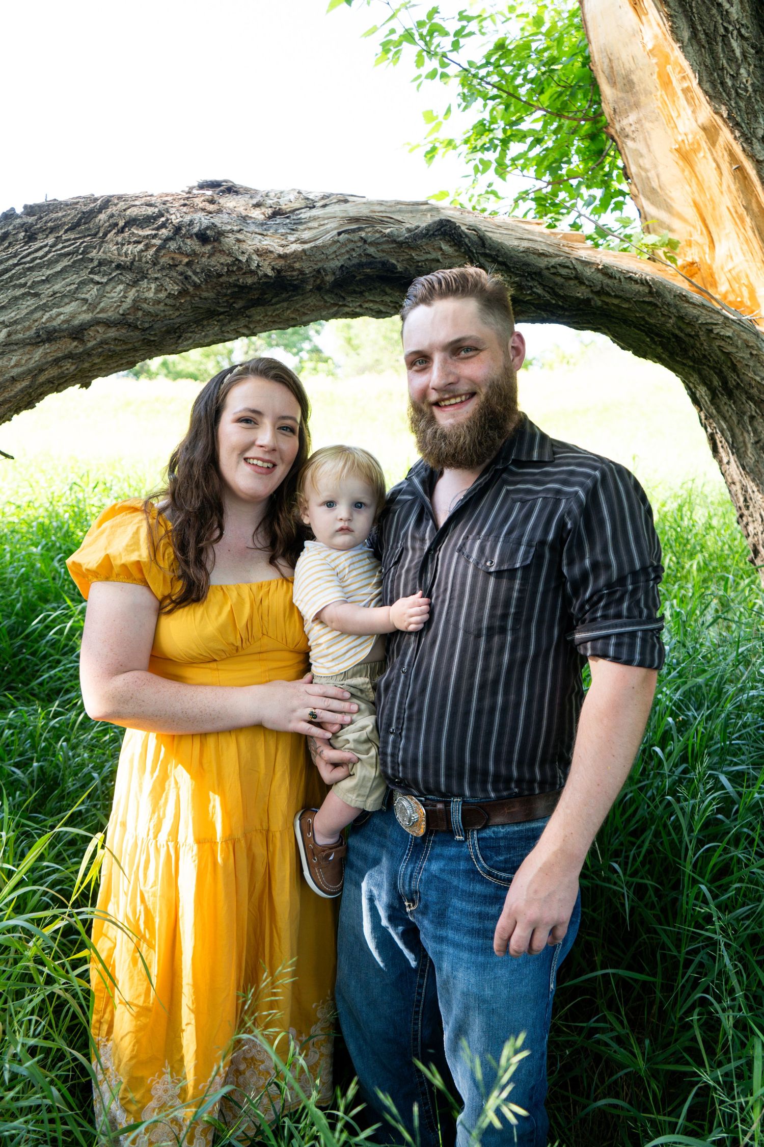 Young family with toddler poses under curved tree branch in lush green outdoor setting.