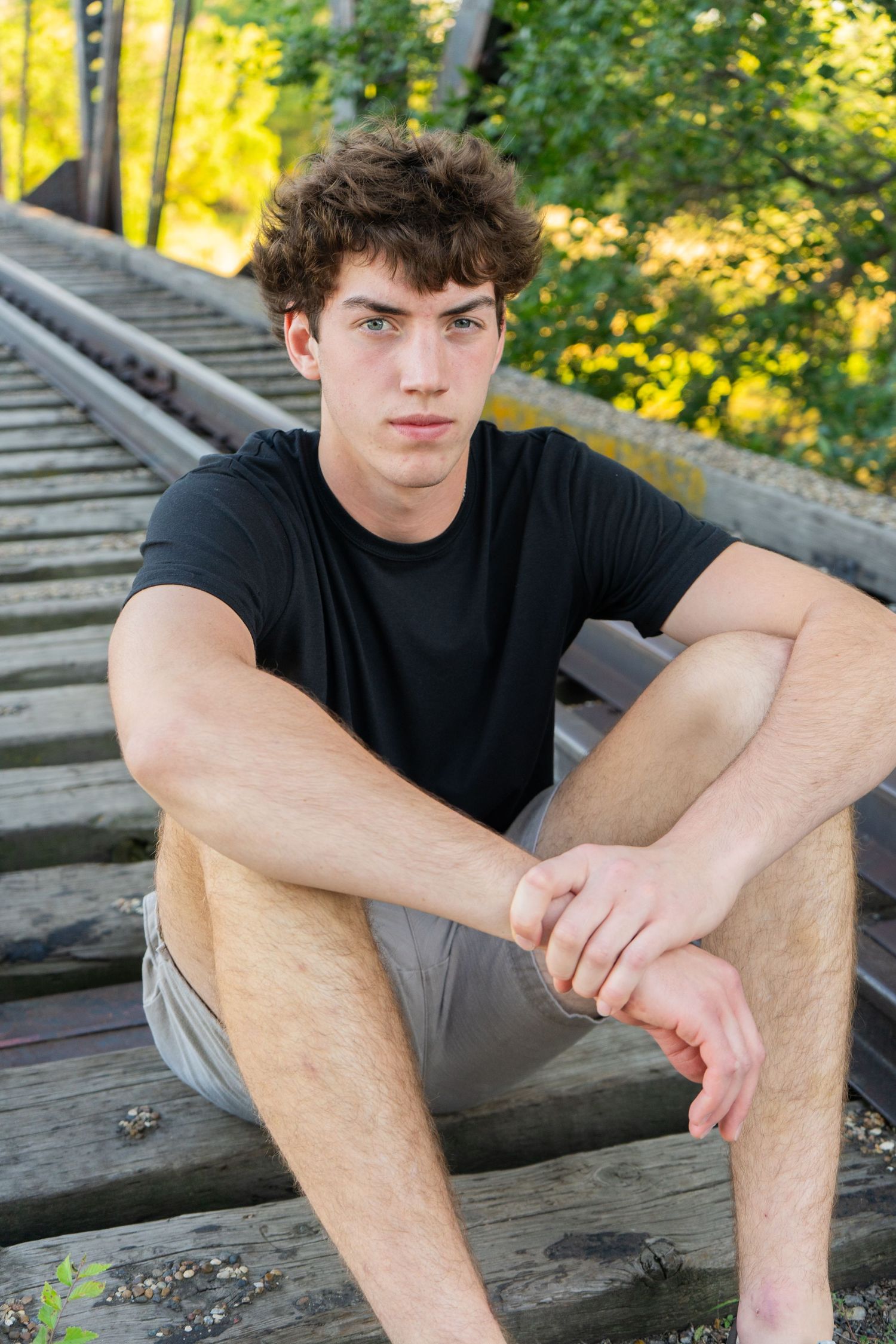 Teenage boy in black t-shirt sitting on wooden railroad tracks surrounded by greenery.