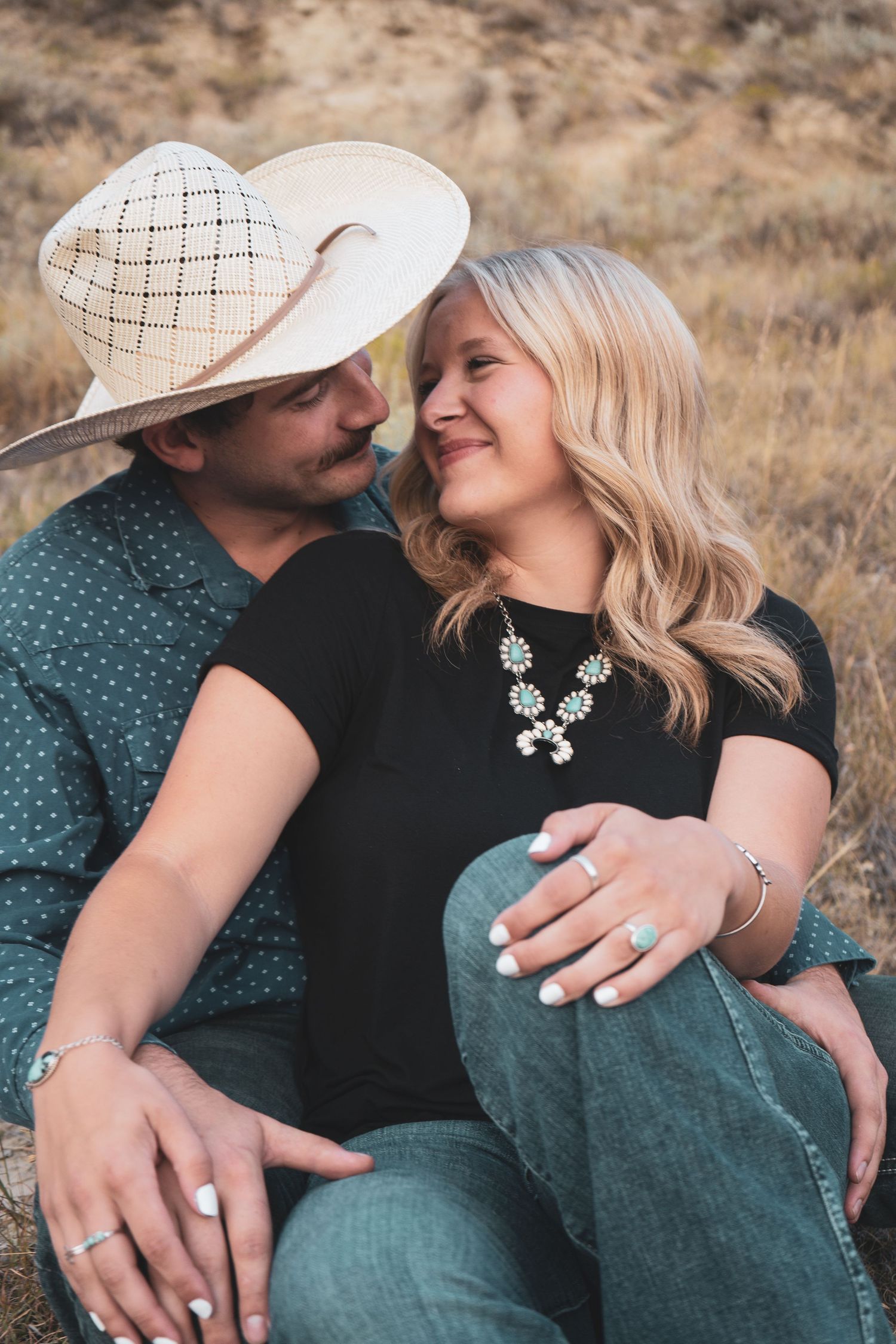 A couple sits closely together outdoors, smiling at each other in a dry desert landscape.