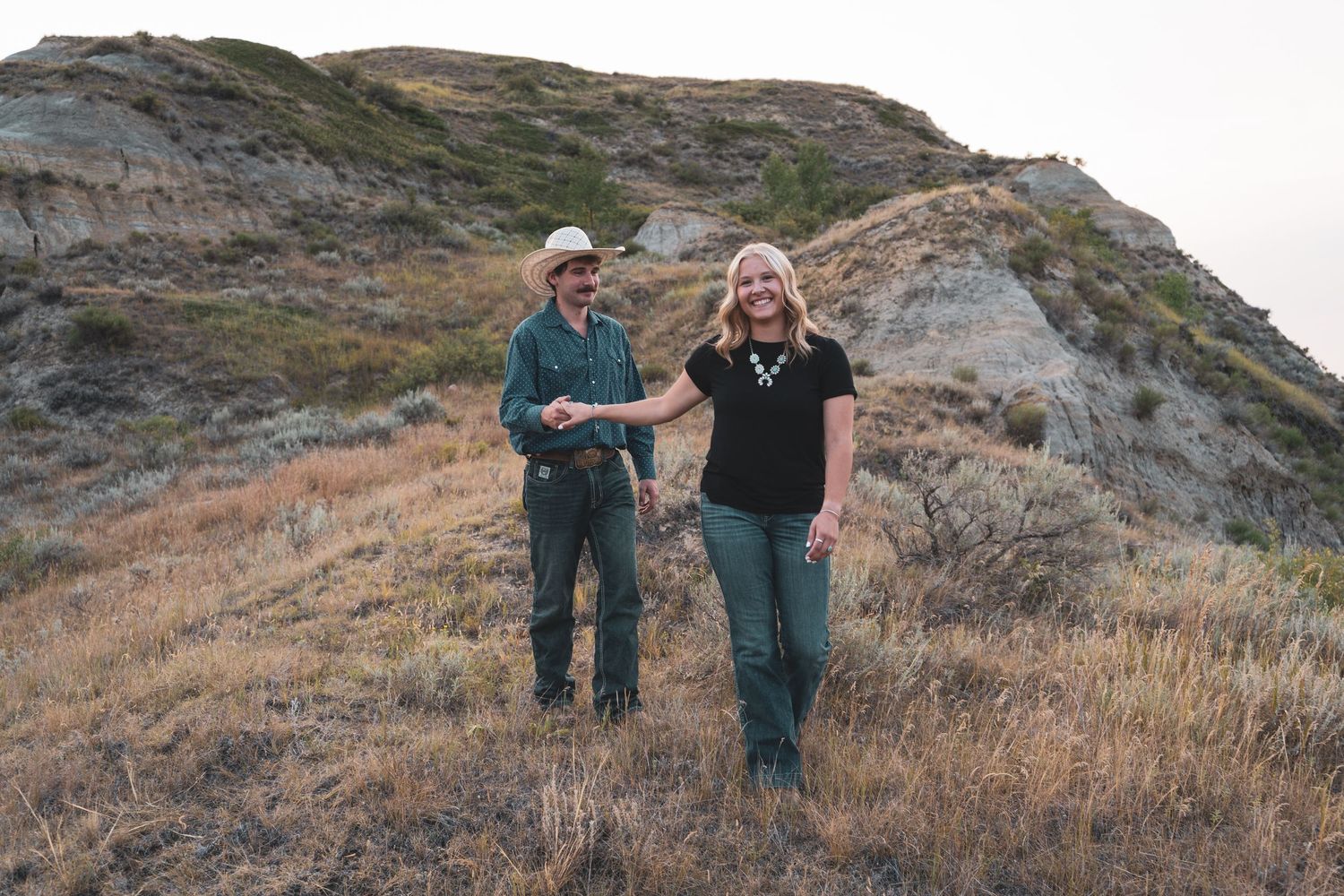 A couple holds hands and walks together on a hillside with badlands scenery in the background.