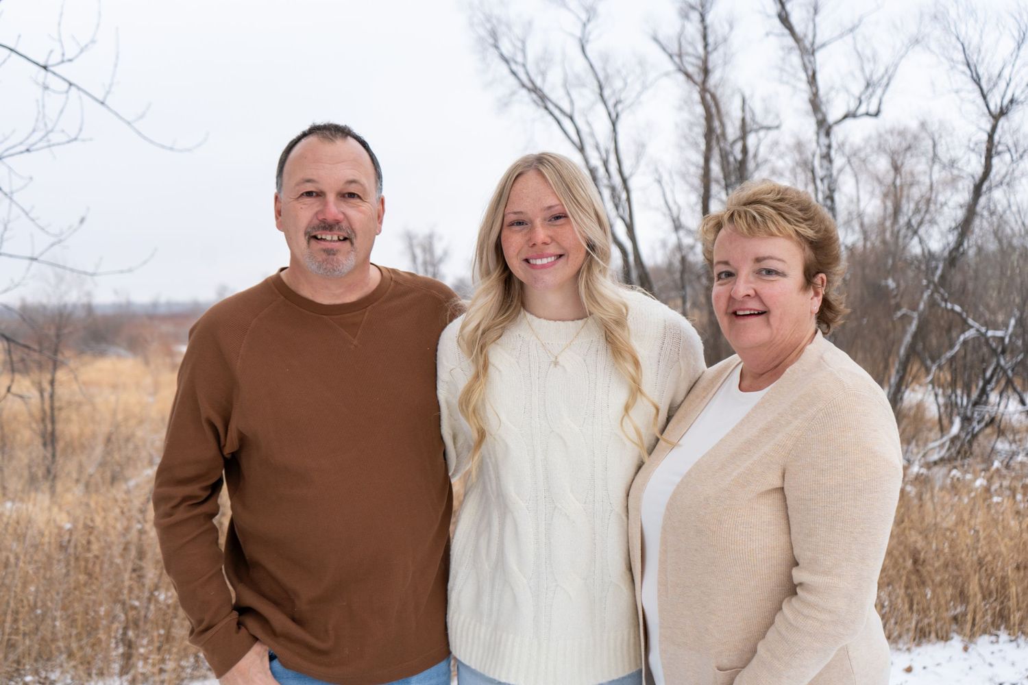 Smiling family of three posing together in snowy outdoor winter landscape with bare trees.