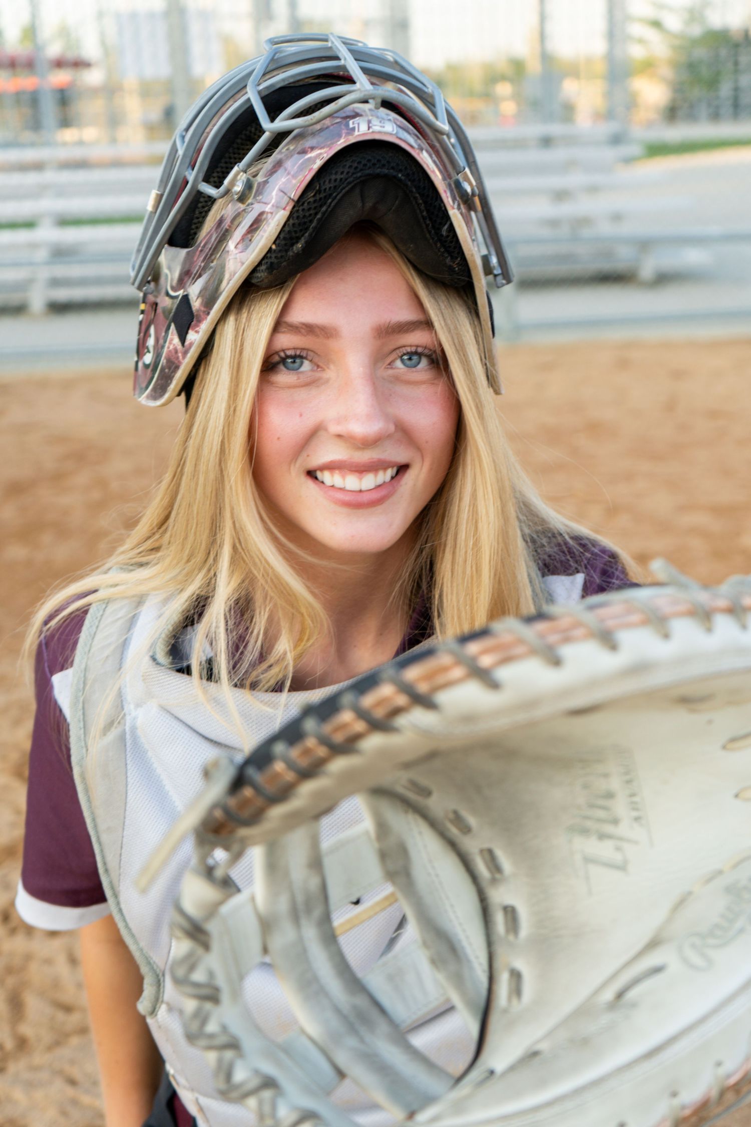 Smiling blonde softball catcher wearing helmet and chest protector holding a catcher mitt close-up.
