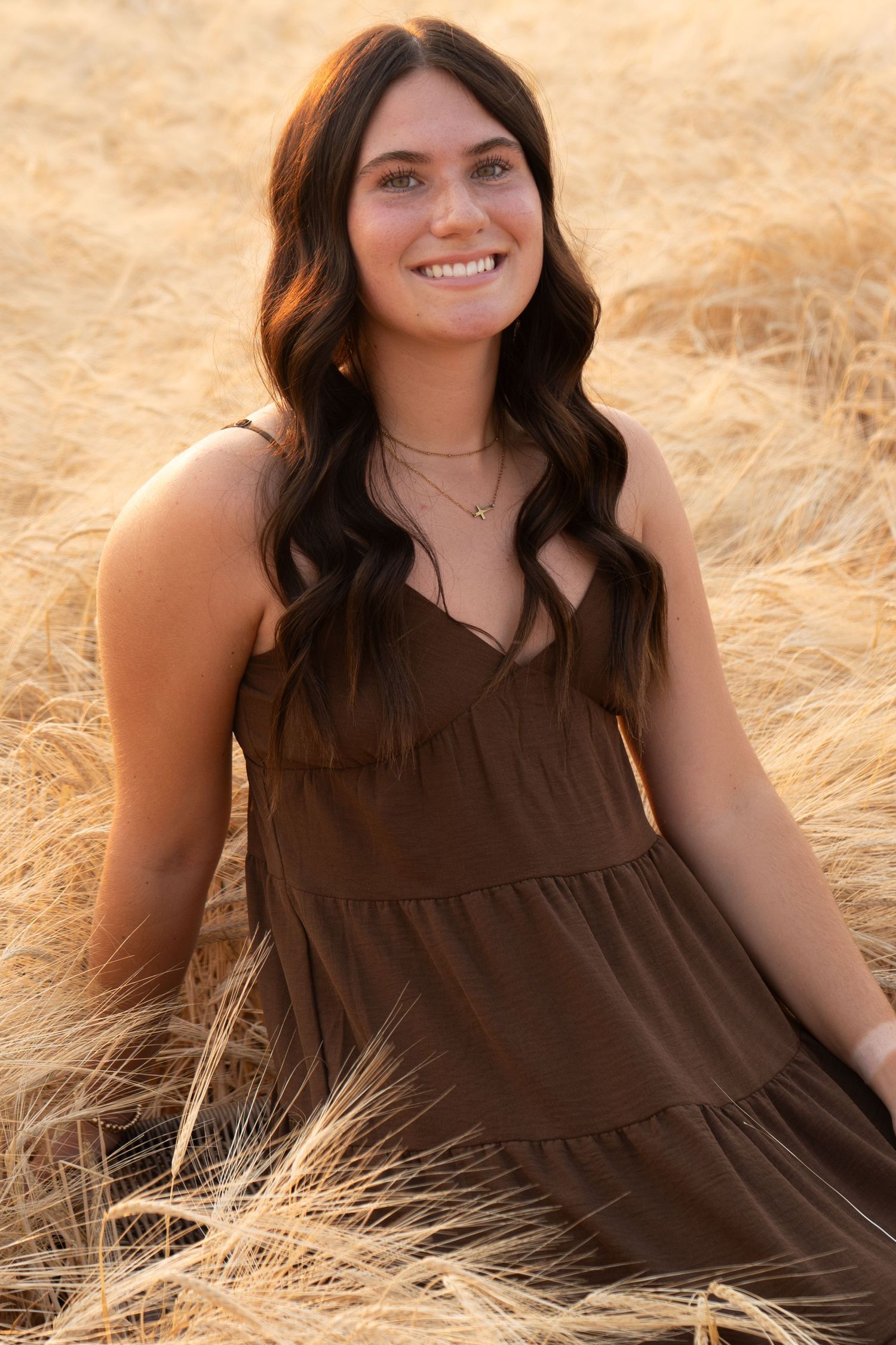 Brunette young woman in brown sundress sits smiling in golden wheat field at sunset.