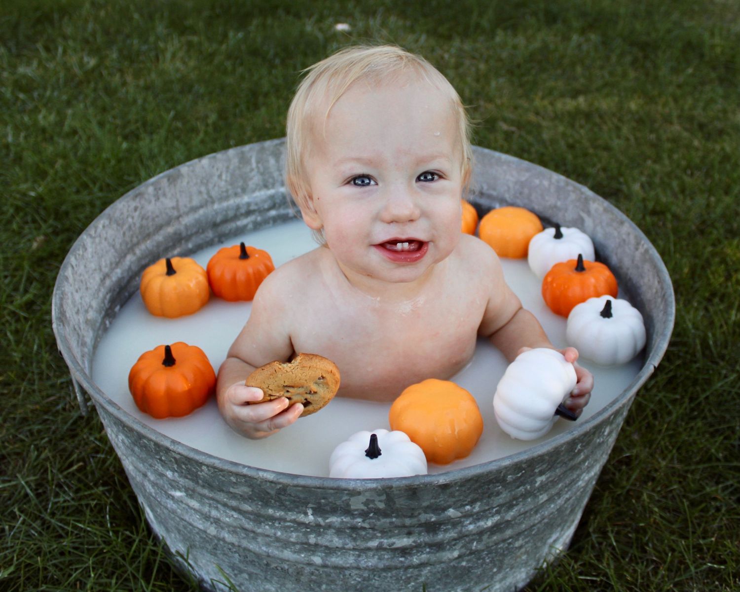 Happy baby smiling in outdoor milk bath with orange and white mini pumpkins in galvanized metal tub.
