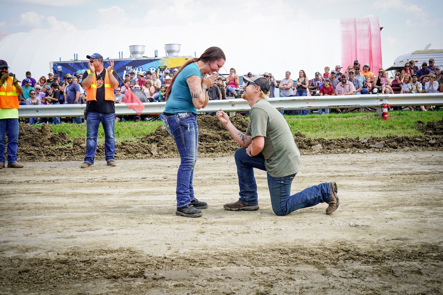 Man proposes to woman on muddy track at outdoor event with crowd watching.