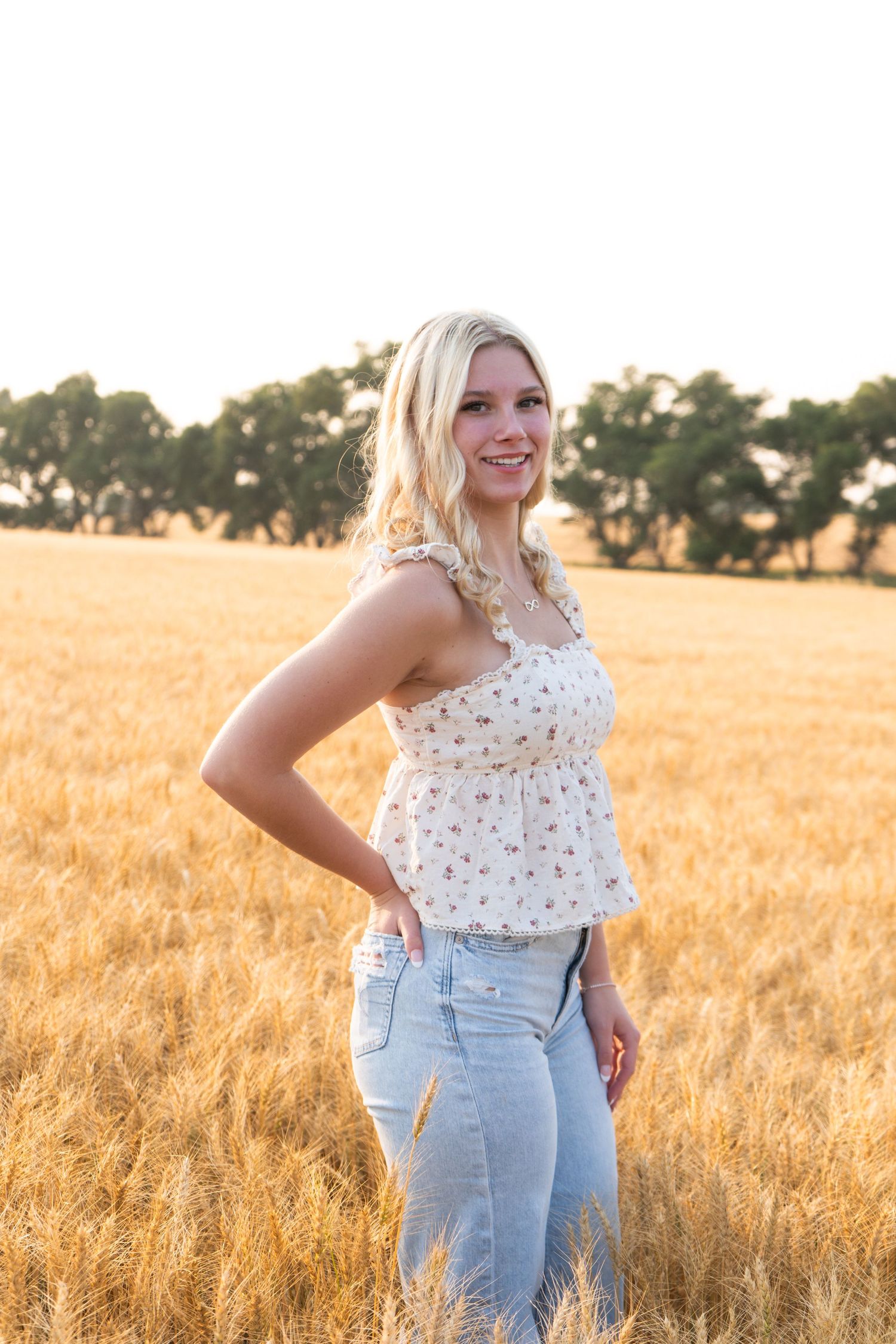 Blonde teenage girl in floral top and jeans smiling in golden wheat field at sunset.