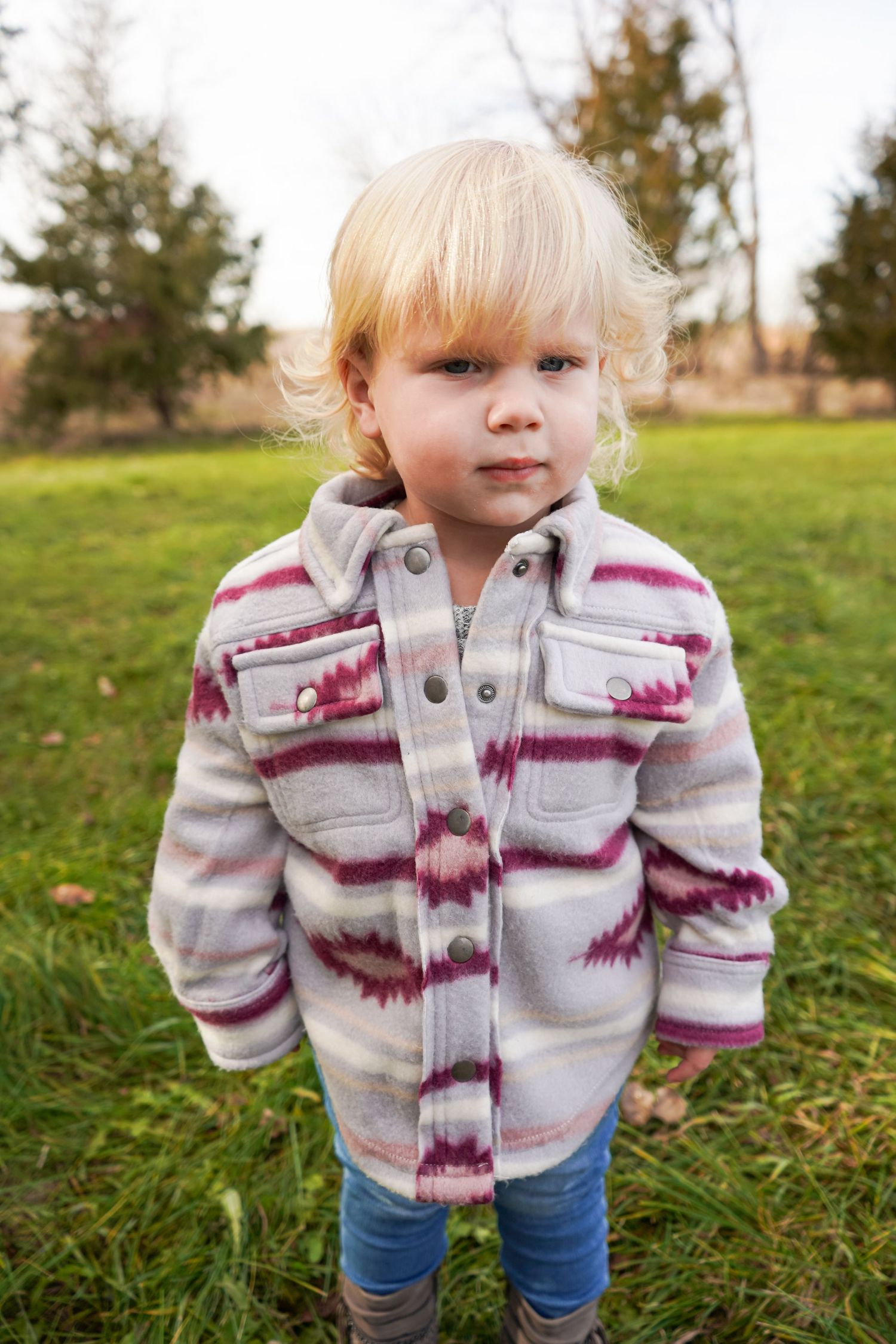 Serious-faced toddler girl with curly blonde hair stands outdoors in a pink and gray fleece jacket.