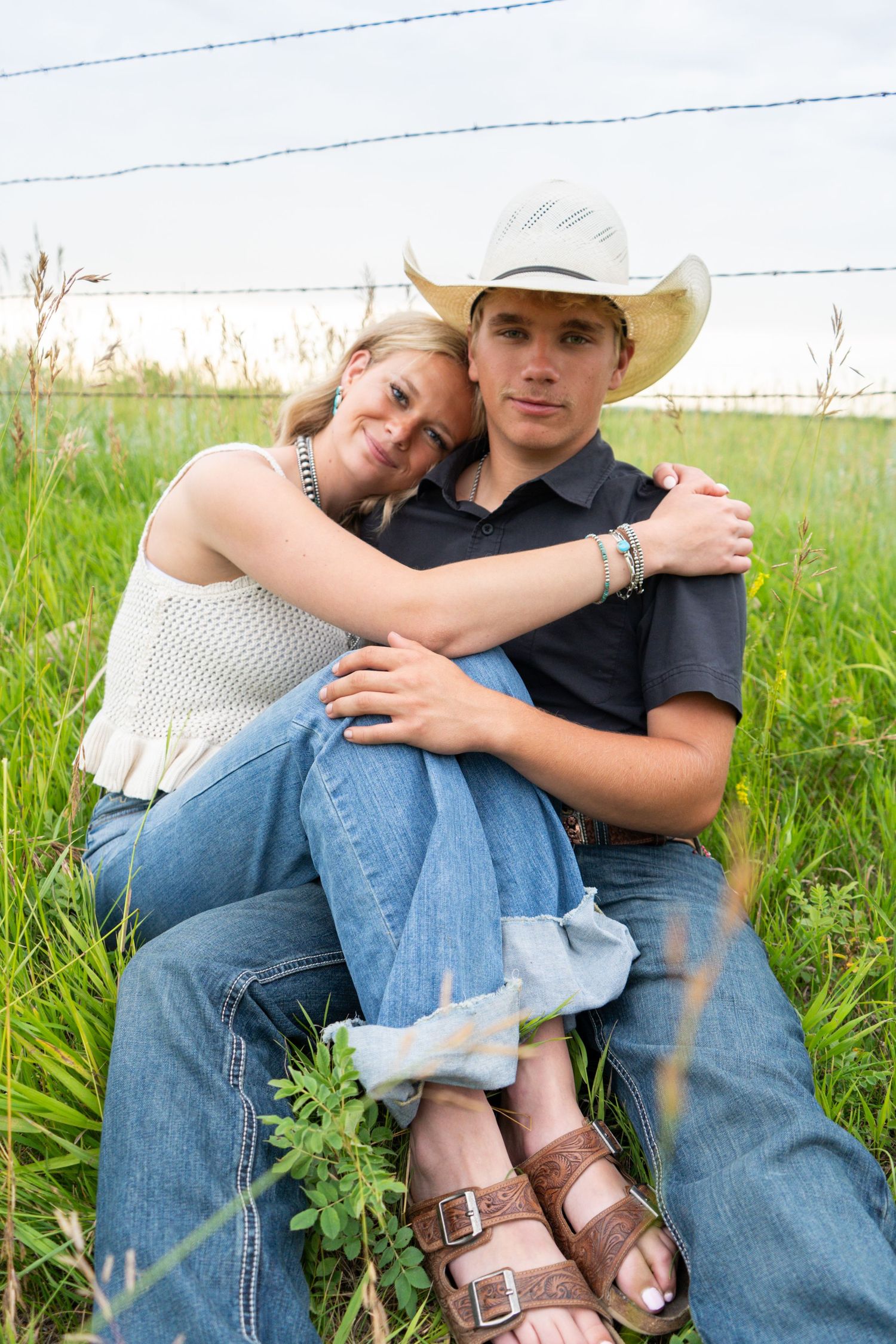 Young couple sitting together in green grass by a barbed wire fence, woman leaning on mans shoulder.