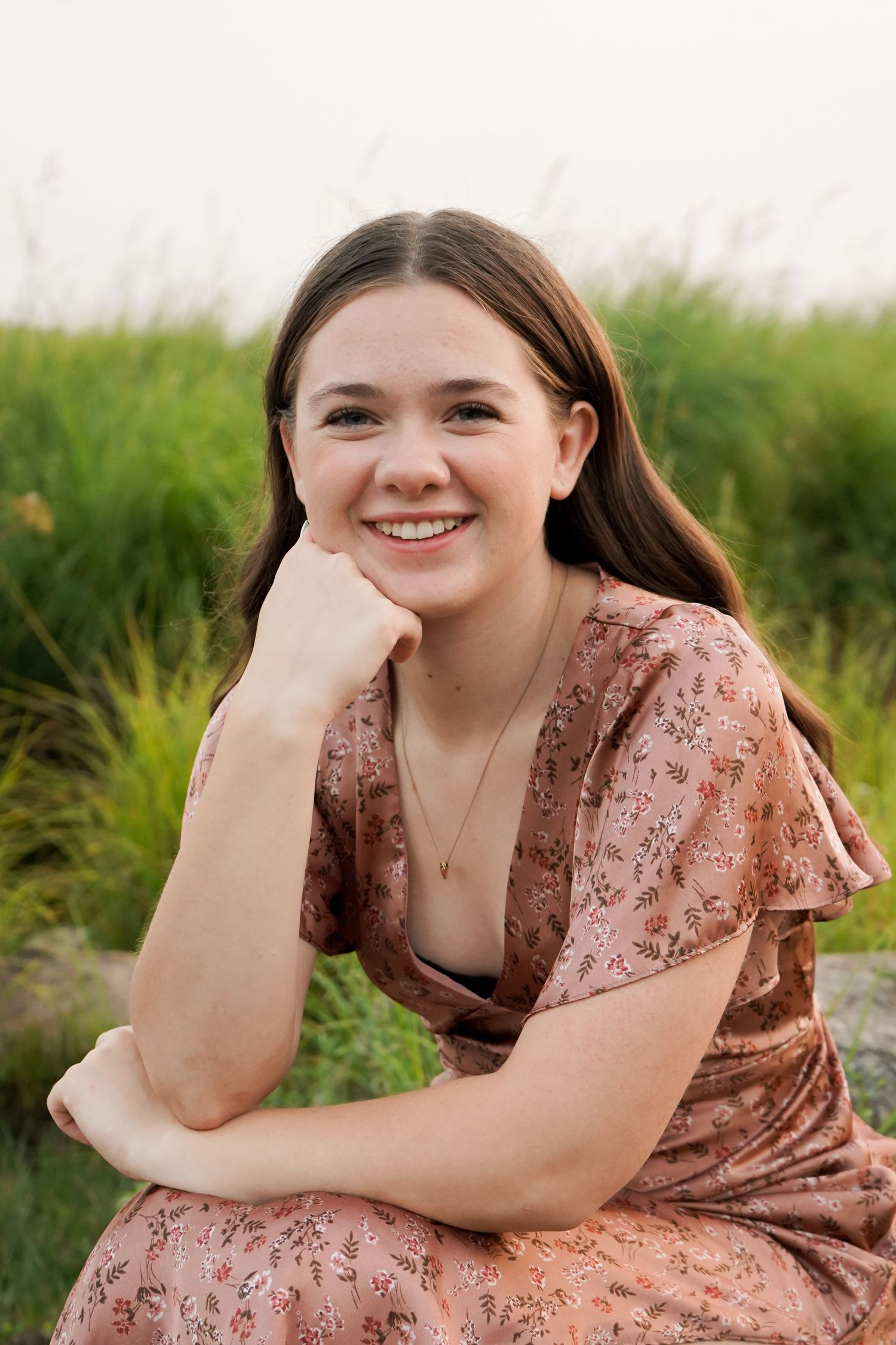 Smiling brunette teen girl in pink floral dress resting chin on hand outdoors.