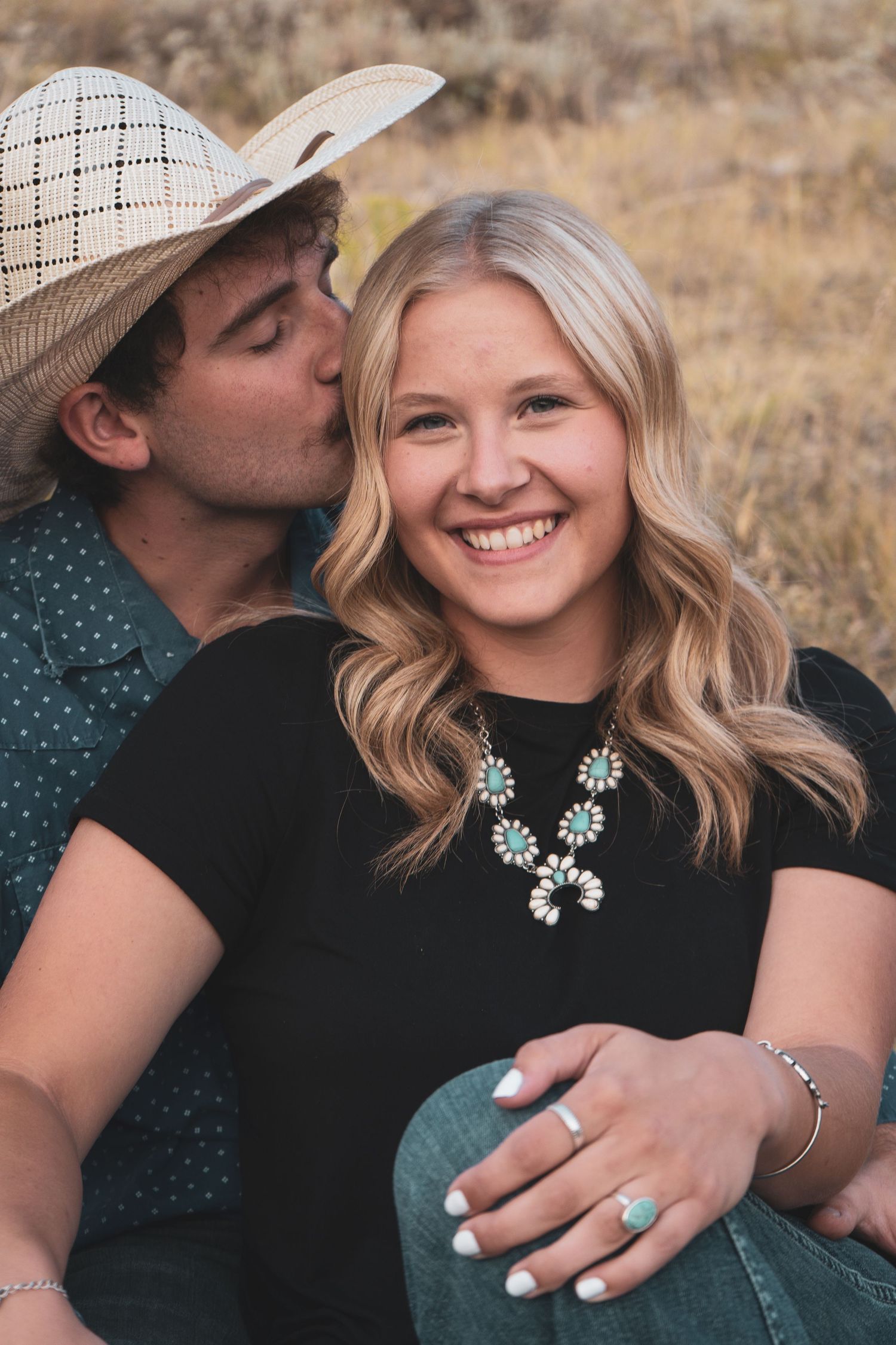 A smiling blonde woman wearing a turquoise necklace sits with a man in a straw hat kissing her cheek.