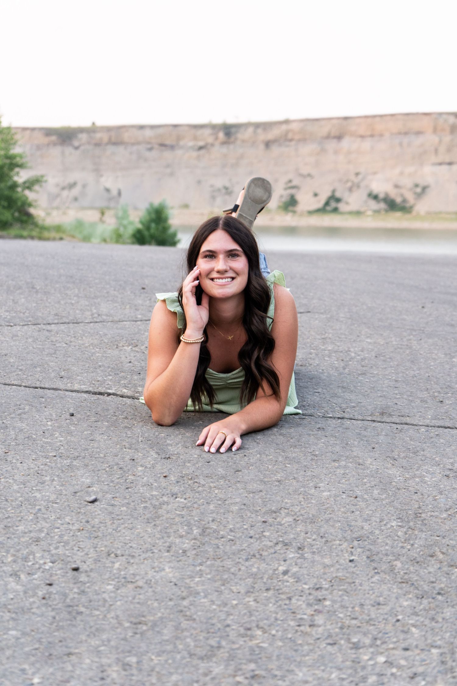 Smiling brunette teen in sage green dress lies on pavement with rocky bluffs in background.