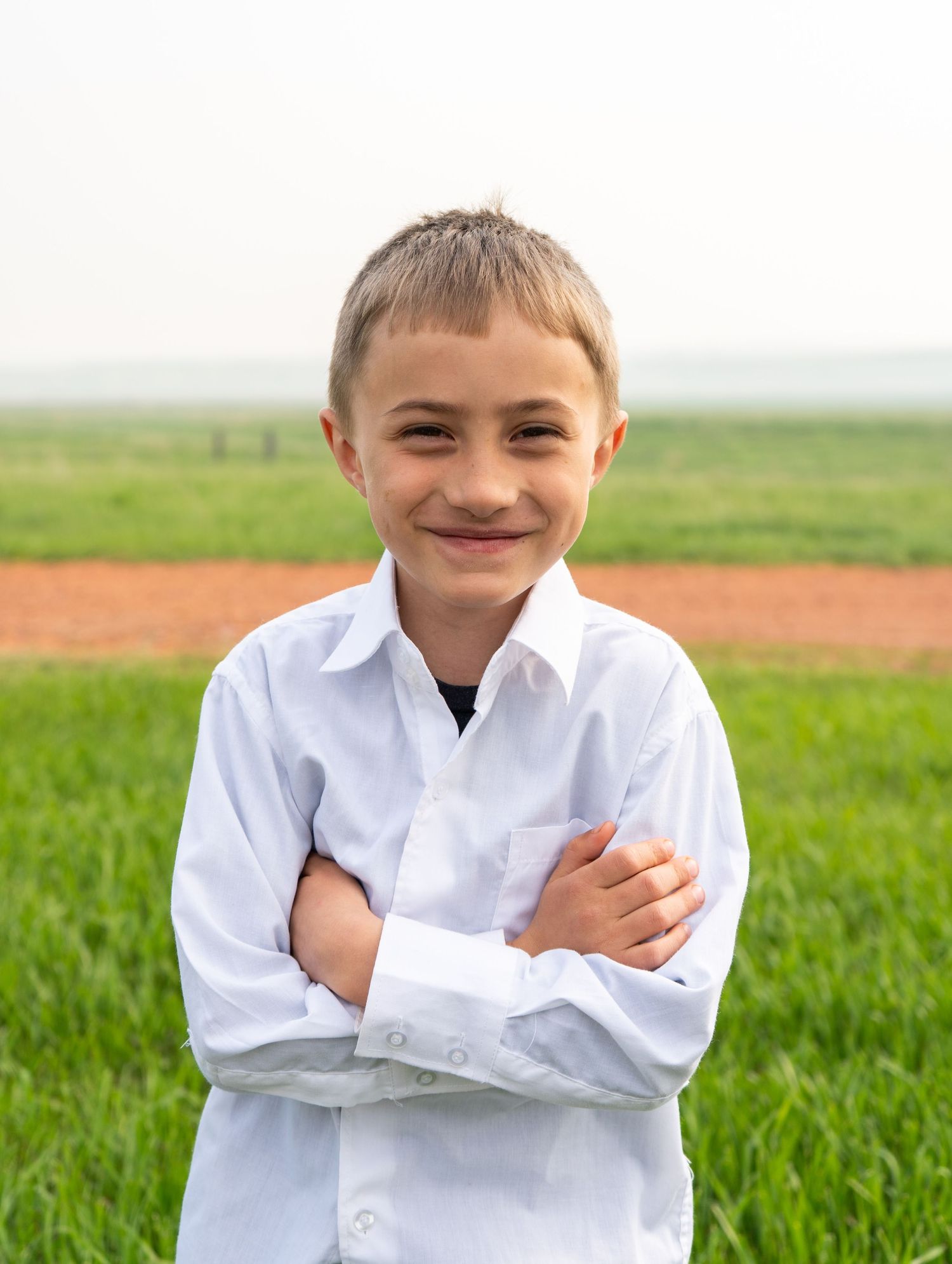 Young boy in white dress shirt standing with arms crossed smiling outdoors.