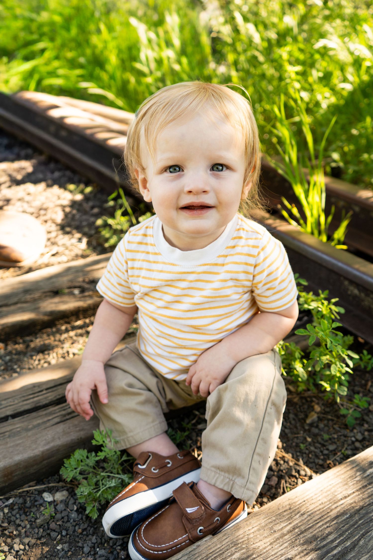 Smiling blond toddler boy sitting on wooden railroad ties outdoors in a yellow striped shirt and brown boat shoes.