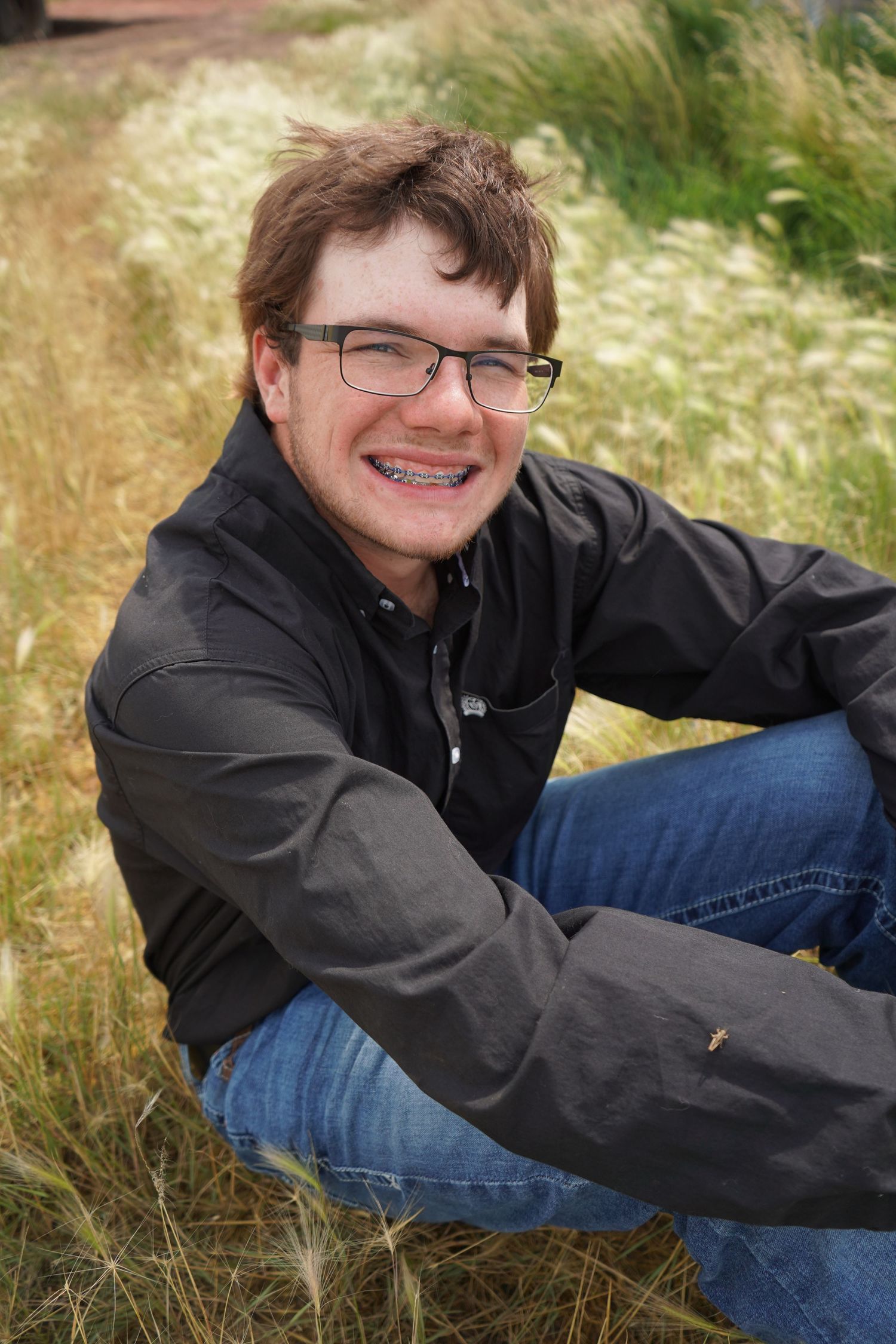 Teenage boy with glasses and braces smiling while sitting in tall grass field.