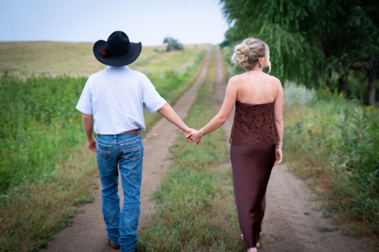 Couple holding hands walking down a country dirt path, man in cowboy hat and woman in brown dress.
