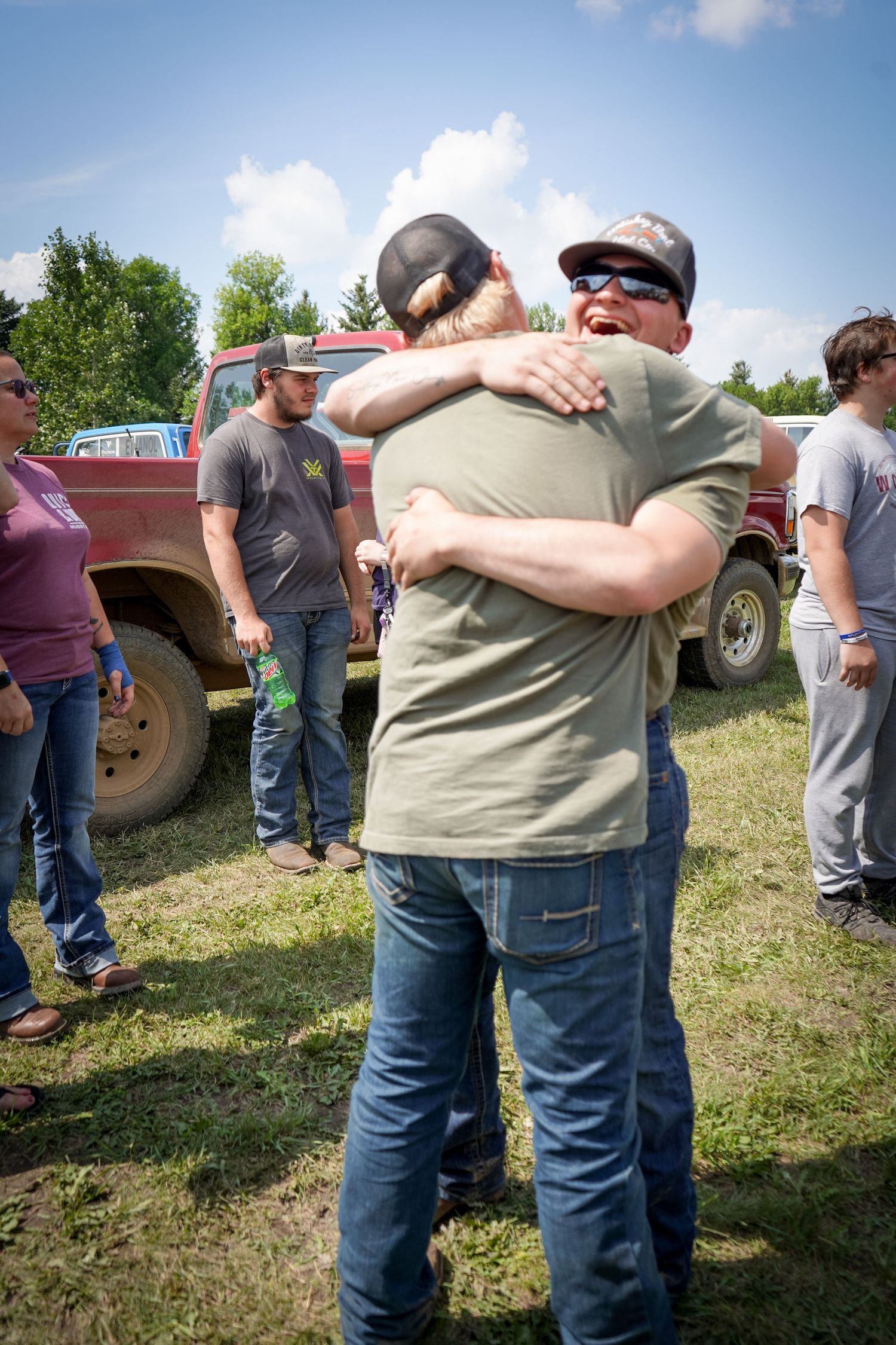 Two men share a warm embrace outdoors near trucks at casual gathering.