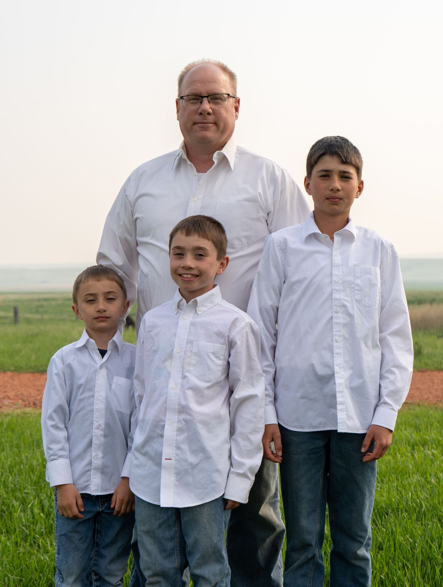 Father posing with three sons in matching white button-up shirts in a green countryside field.