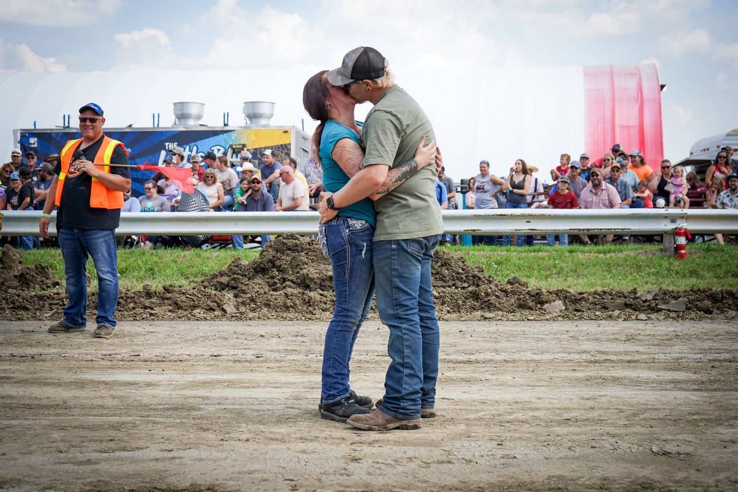 Newly engaged couple shares a kiss on dirt track with crowd behind them.